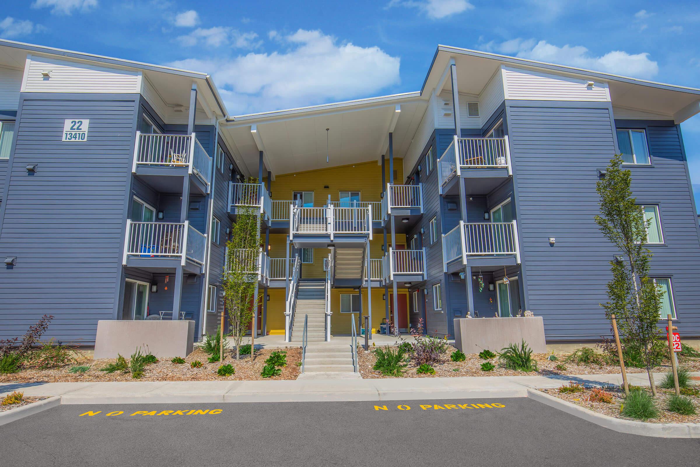 Modern apartment building with three stories, featuring blue and yellow exterior colors. The front area includes landscaped greenery, parking spaces labeled "N O PARKING," and balconies on the upper floors. The sky is partly cloudy.