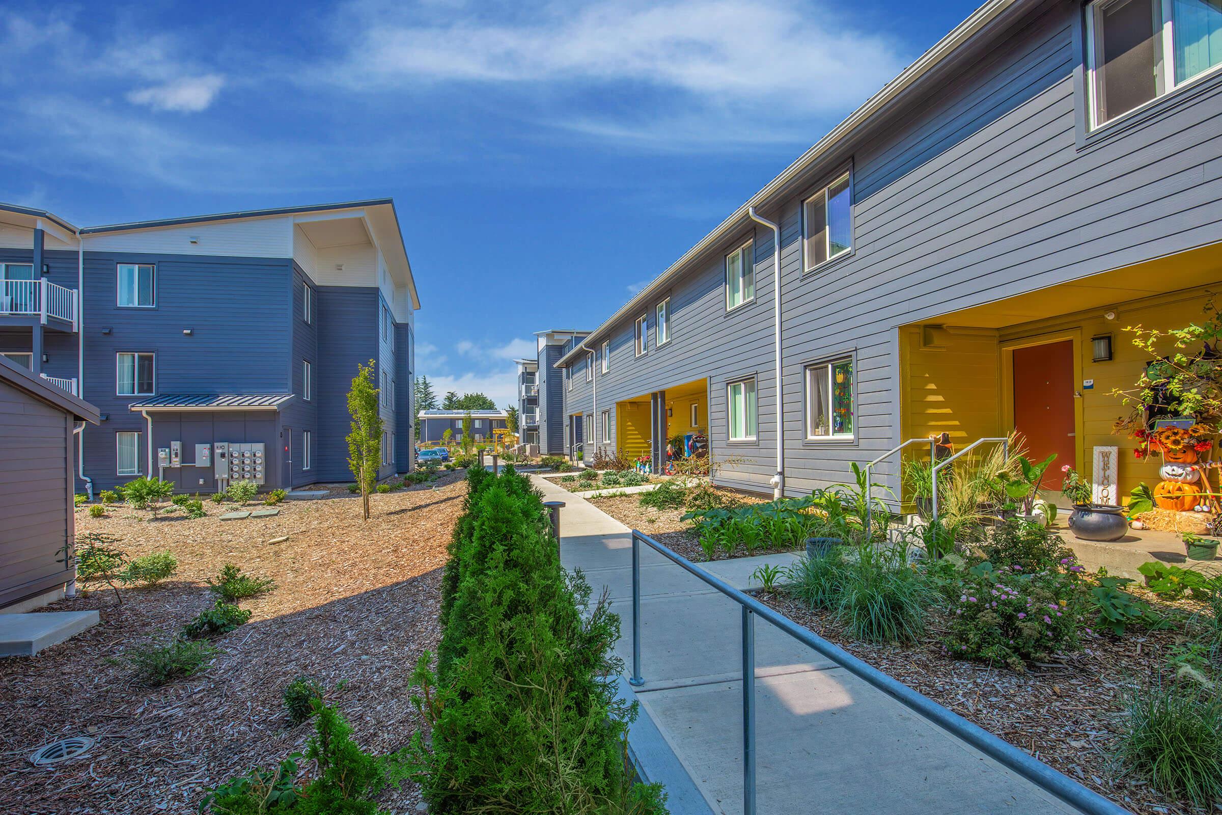 A well-maintained apartment complex with a landscaped pathway. Colorful exteriors of buildings are visible, along with green plants and shrubs lining the walkway. The sky above is bright blue with a few clouds, creating a welcoming and sunny atmosphere.