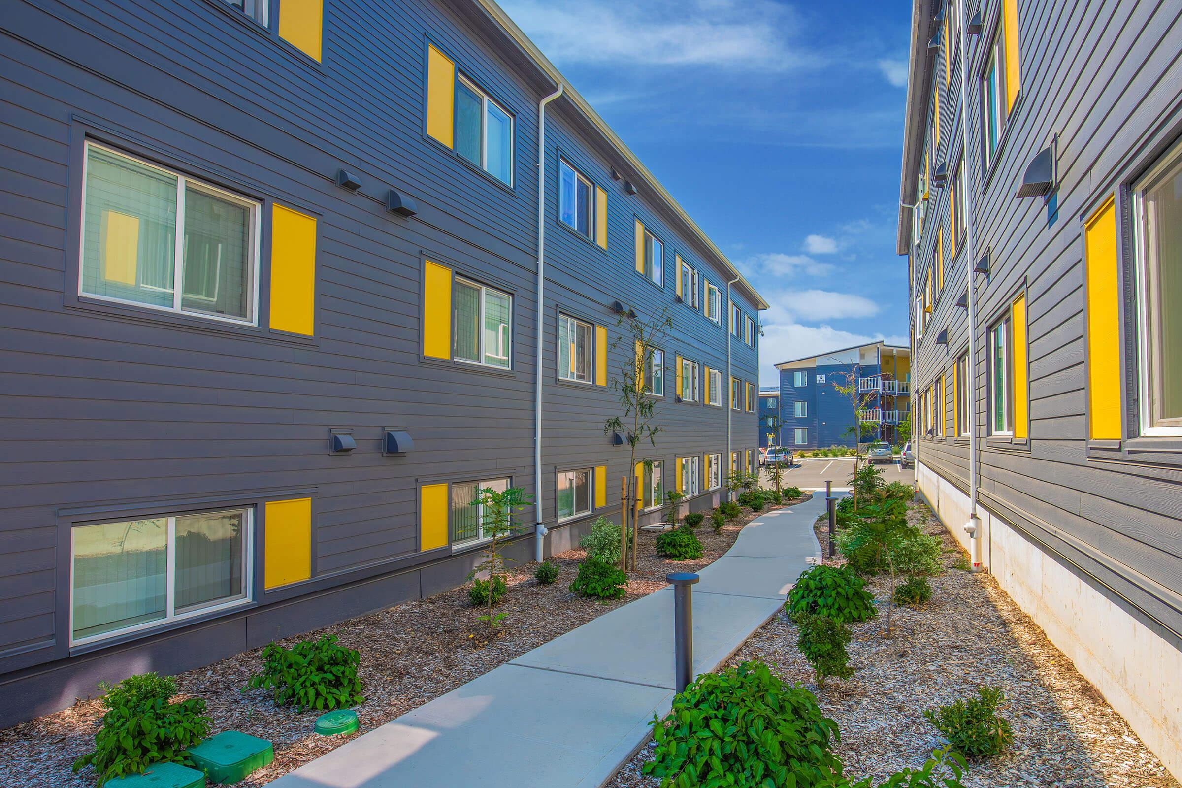 A modern apartment complex with two buildings featuring gray exteriors and yellow window trim. A paved walkway runs between the buildings, bordered by landscaped areas with small plants and gravel. The sky is clear with some clouds, creating a bright, inviting atmosphere.