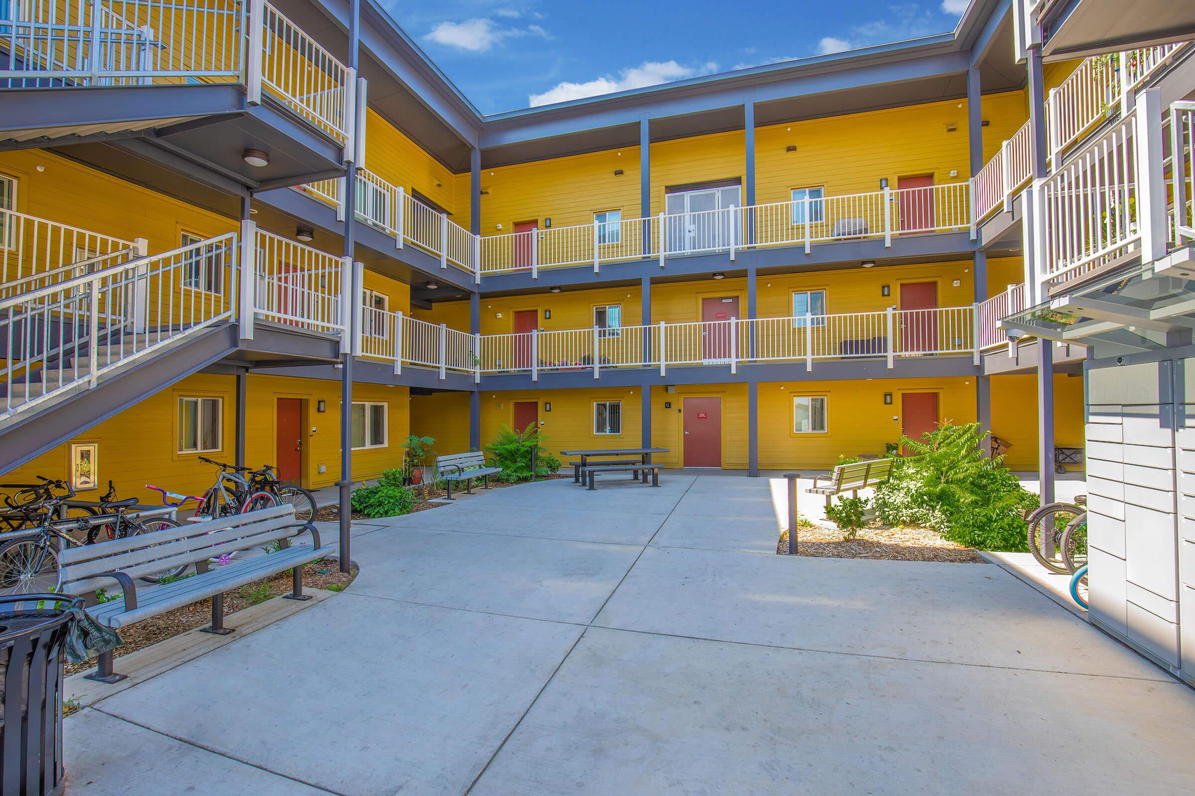 An outdoor courtyard of a modern, multi-story apartment building featuring bright yellow walls. There are several doorways leading to apartments, benches, a picnic table, and bicycles parked in the area. The scene is sunny with a clear blue sky and greenery around the courtyard.