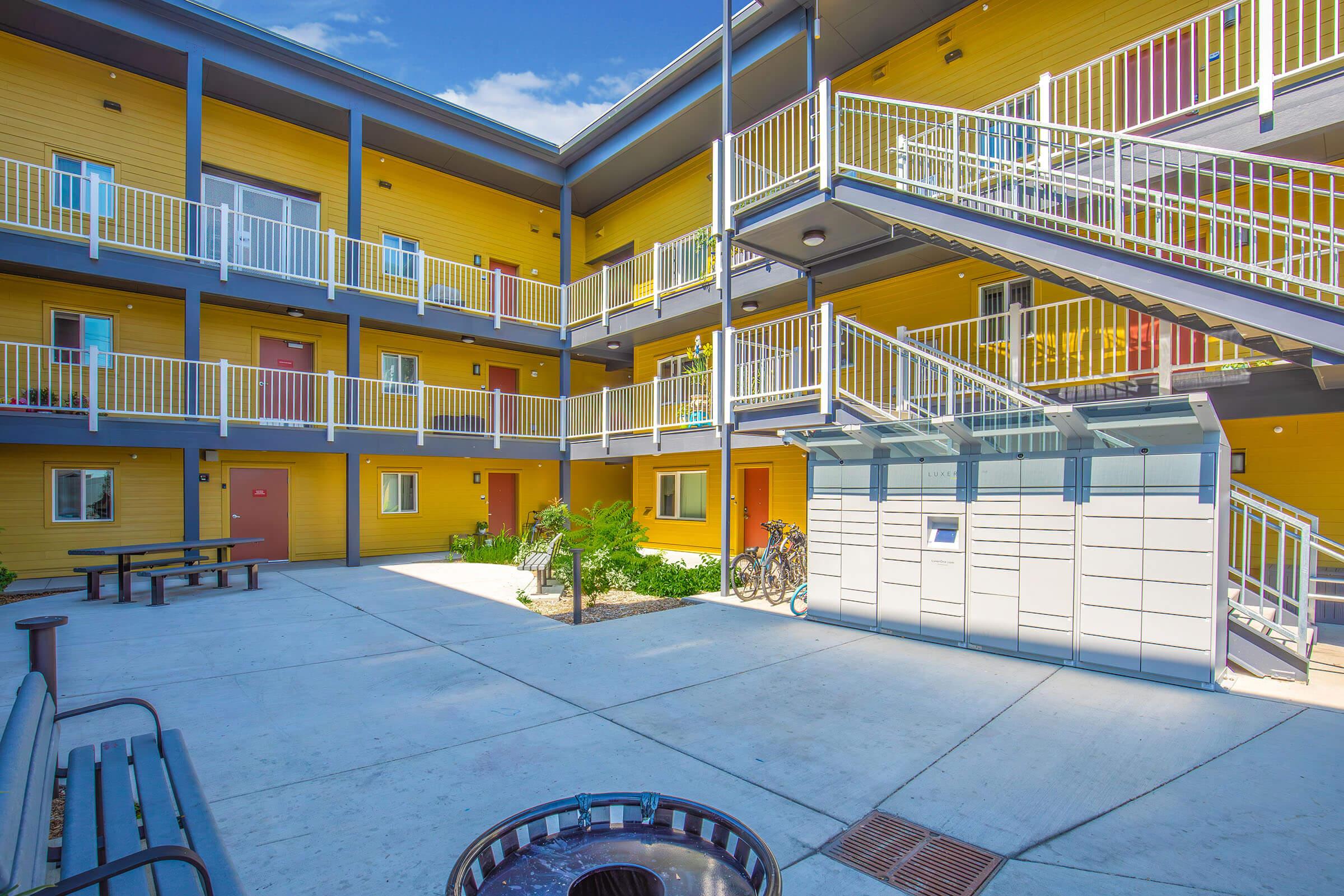 A bright yellow apartment complex featuring multiple levels, with balconies and staircases. The courtyard has benches, bike storage, and well-maintained landscaping. Windows are visible in several units, creating a welcoming outdoor space. The sky is clear and blue, suggesting a sunny day.