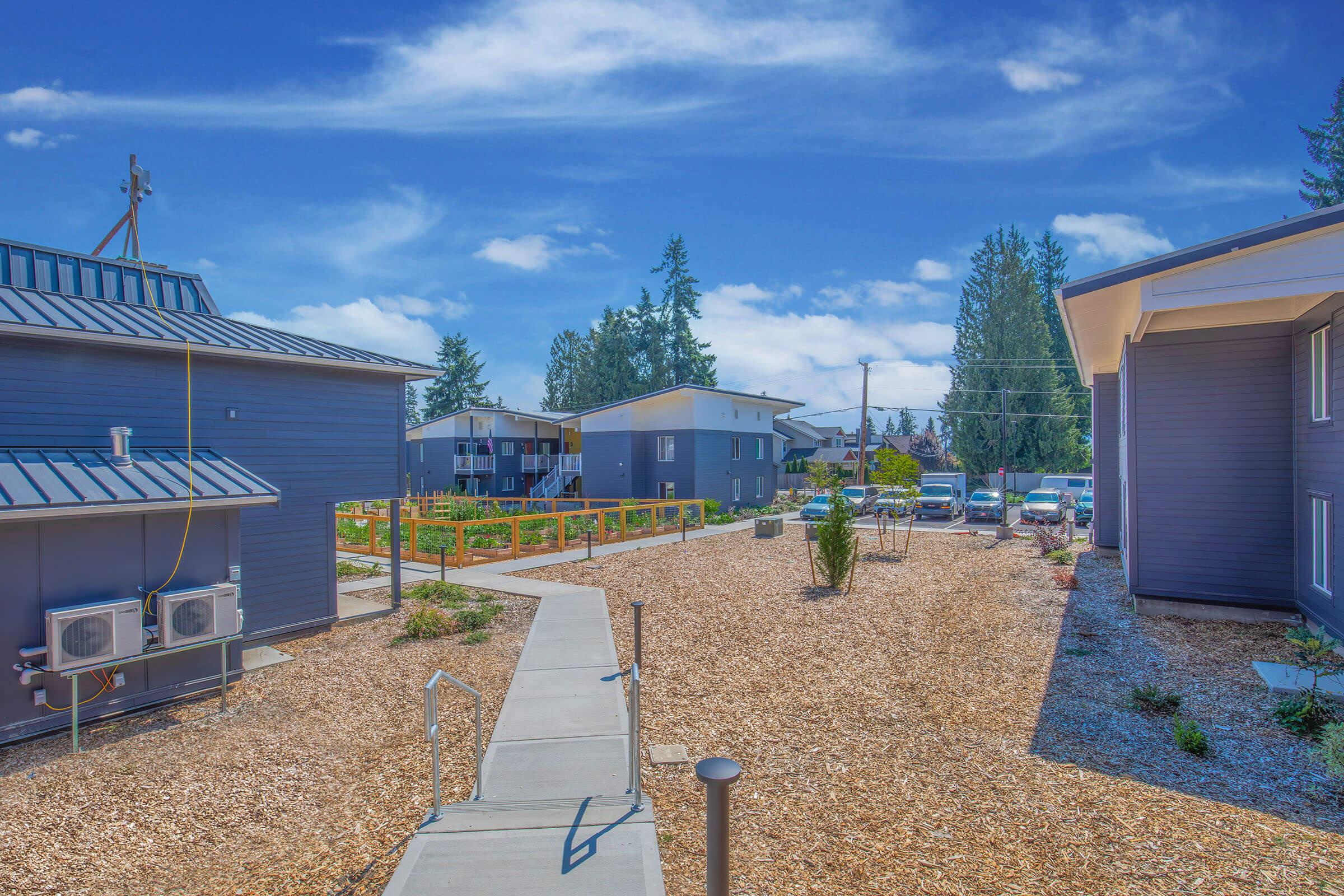 A landscaped residential area featuring modern buildings. The pathway leads through a gravel yard with shrubs and trees, bordered by wooden fences. Cars are parked at the side, and the sky is bright with scattered clouds. The scene reflects a peaceful, well-maintained community environment.