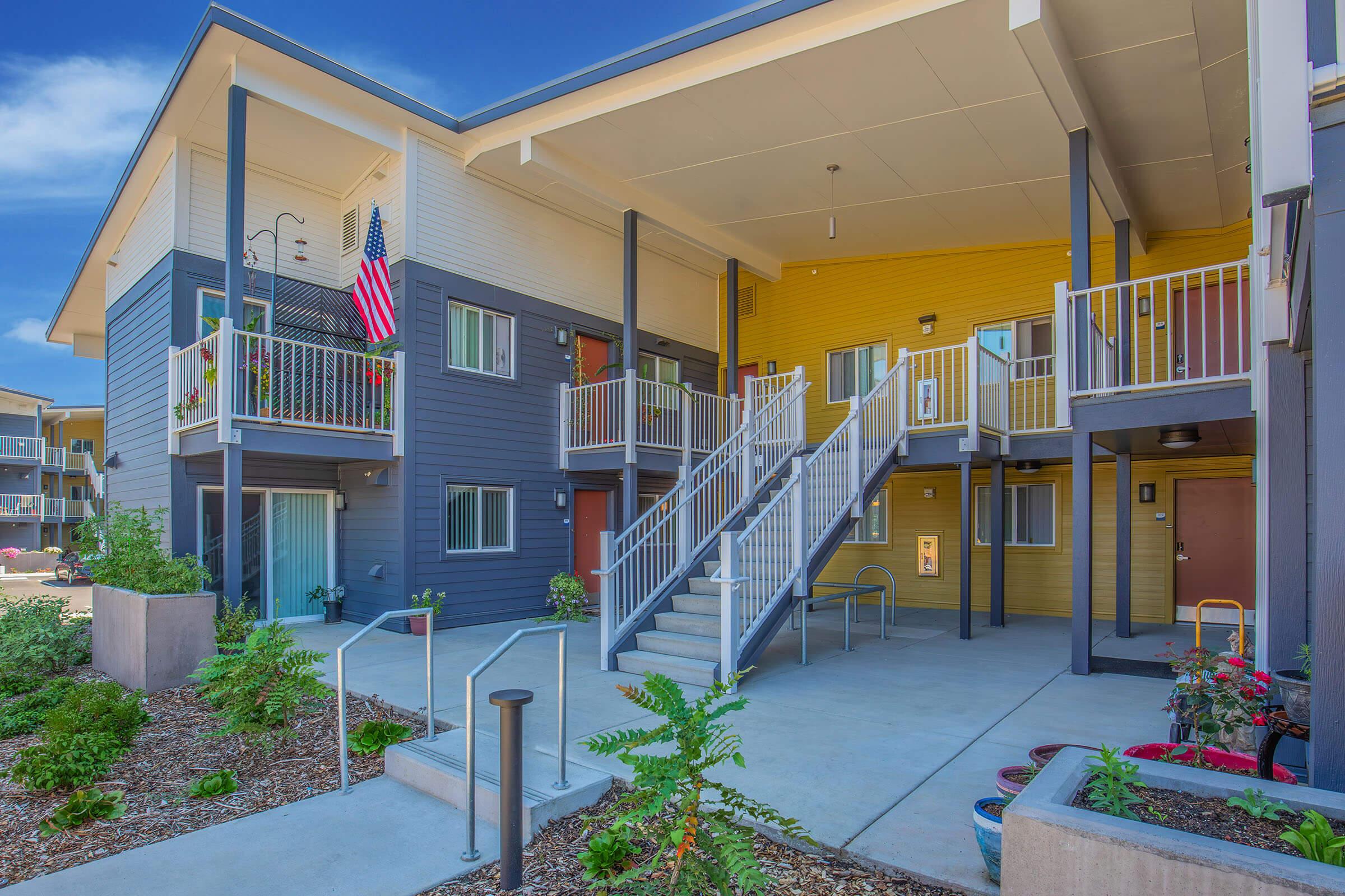 Exterior view of a multi-story apartment building with a mix of blue and yellow siding. Features include a staircase, potted plants, and an American flag displayed on a balcony. The area is well-lit and landscaped, with a modern design.