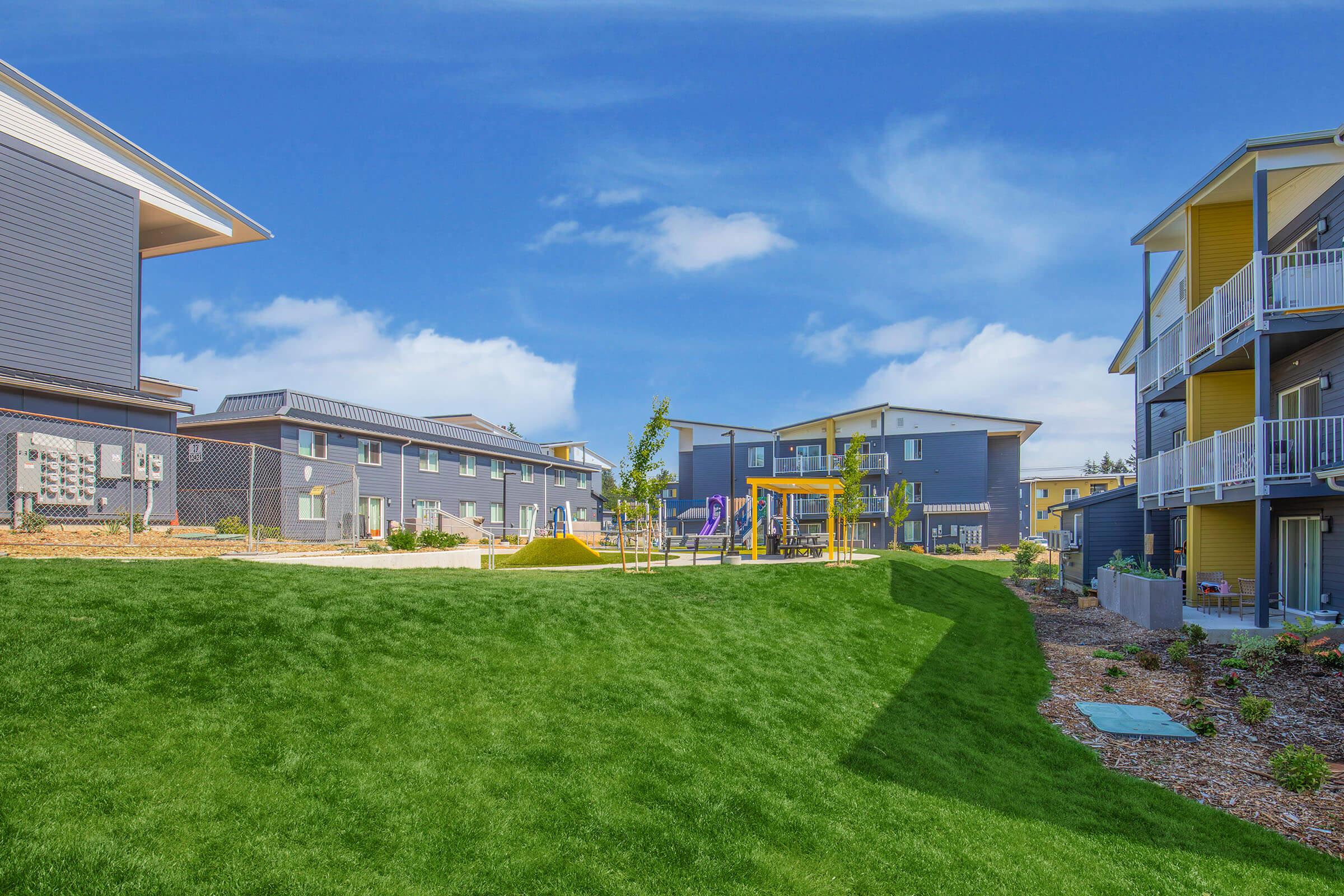 A vibrant residential area featuring modern apartment buildings in shades of blue and yellow, surrounded by manicured green lawns. In the foreground, there is a playground with play structures, and the background shows additional buildings against a clear blue sky.