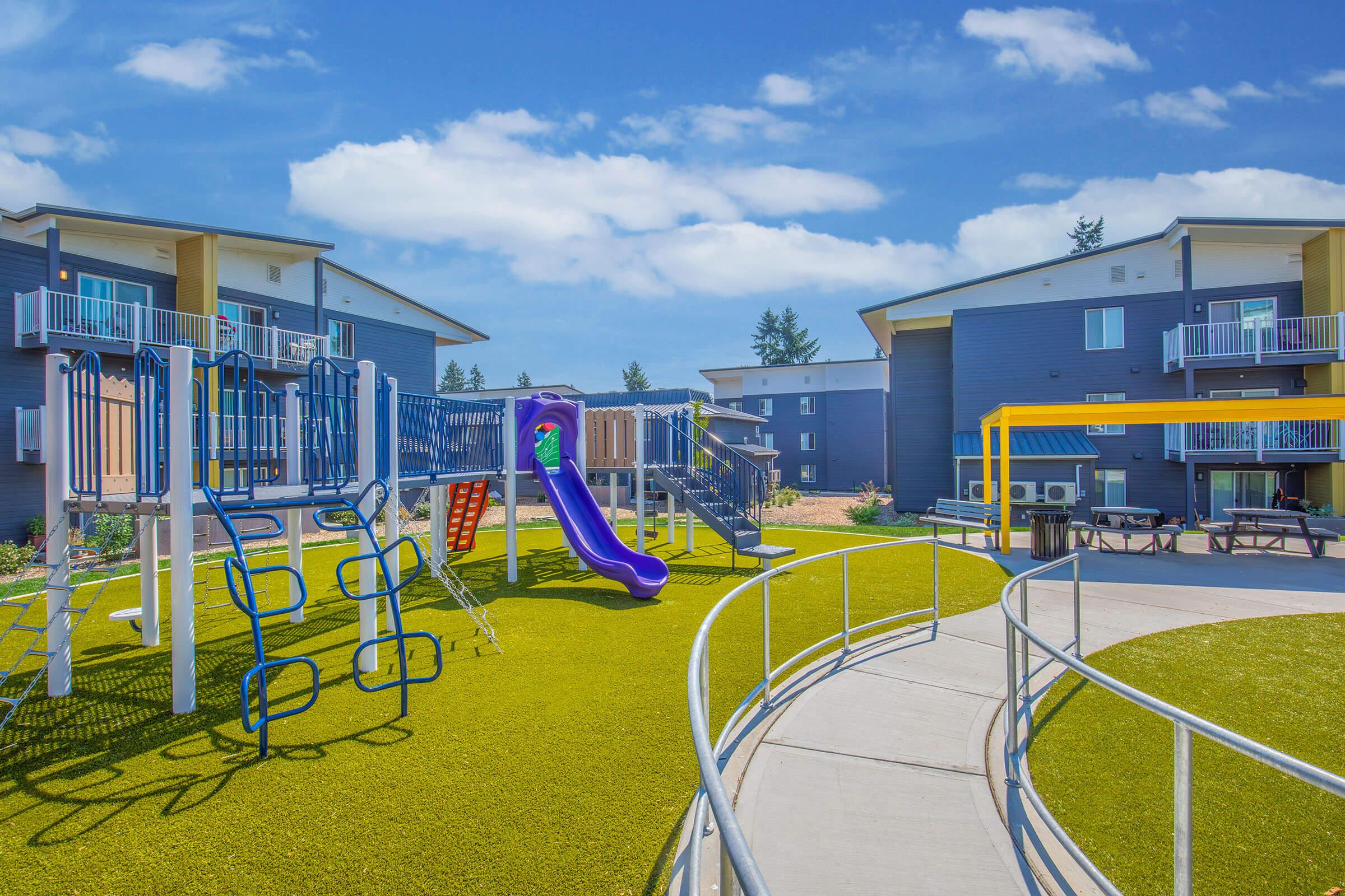 Colorful playground equipment with slides and climbing structures is set against a backdrop of modern apartment buildings. The playground area features green grass and a paved walkway, surrounded by trees and a bright blue sky.