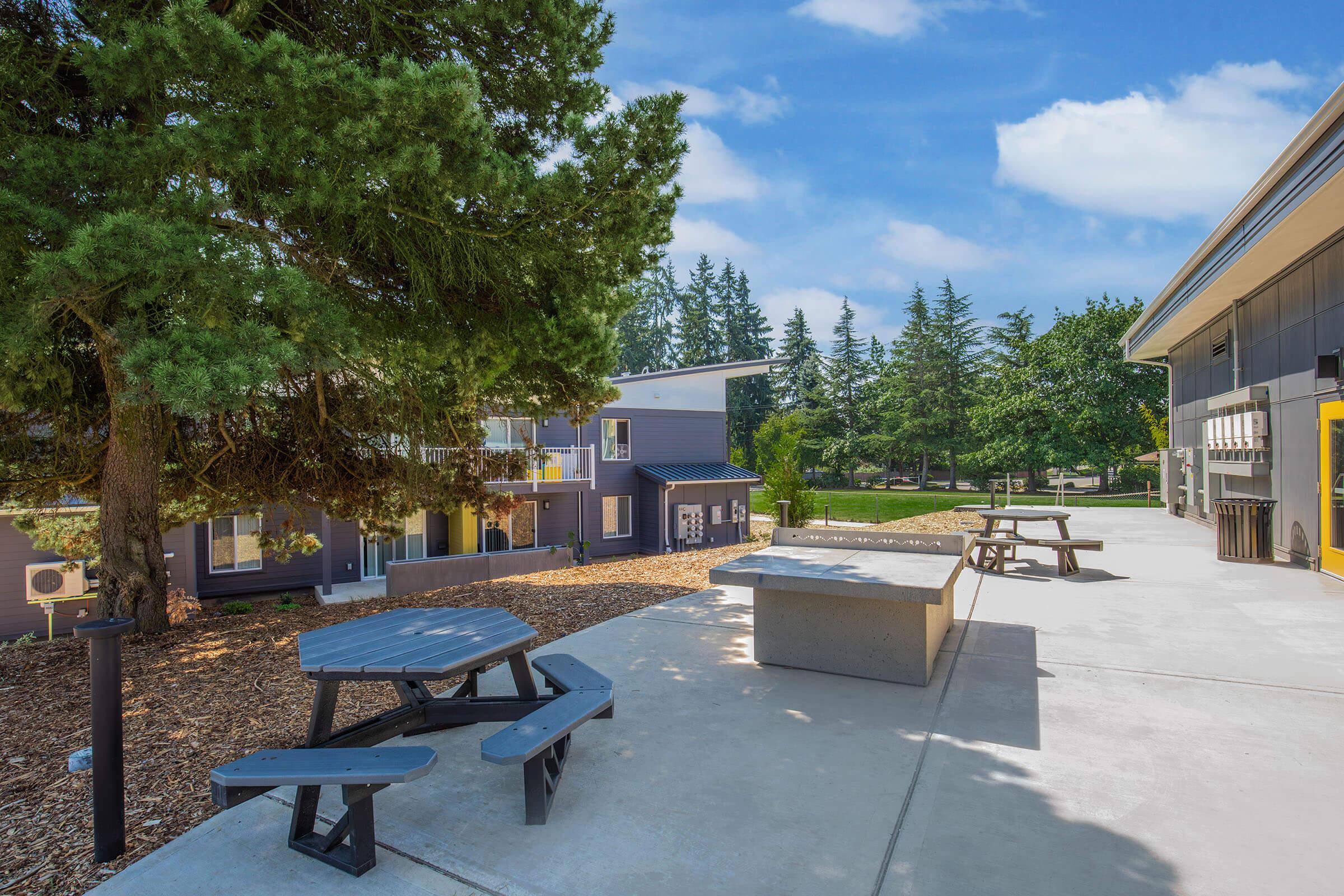 A peaceful outdoor area featuring a picnic table and a concrete bench, surrounded by trees and colorful buildings. The scene is bright and inviting, with a clear blue sky and well-maintained landscaping, creating a perfect spot for relaxation or social gatherings.