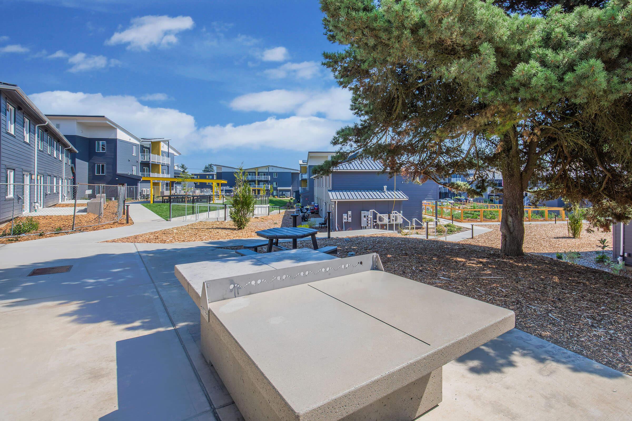 Outdoor area featuring a concrete ping pong table, surrounded by landscaped grounds with trees and buildings in the background. The scene is bright and sunny, with blue skies and a few clouds, showcasing a modern residential complex. Picnic tables are visible nearby, inviting social gatherings.