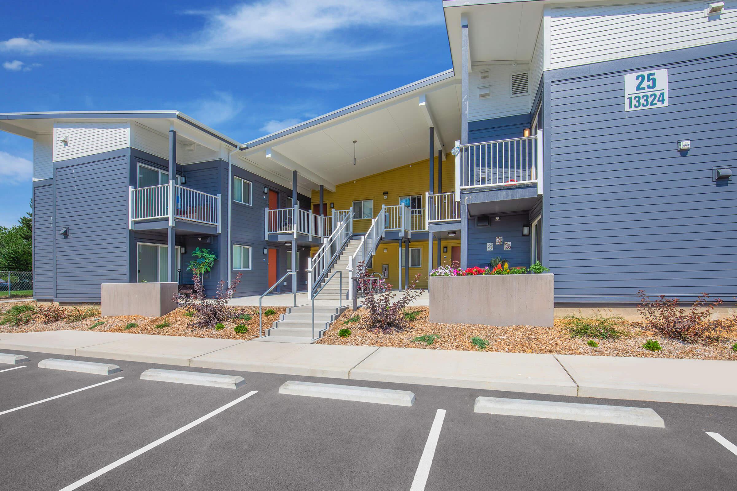 Modern multi-unit apartment building with a two-tone exterior featuring gray and yellow walls. The structure has a covered entrance, multiple balconies, and a landscaped front area with shrubs. A parking lot is visible in the foreground with marked spaces. Clear blue sky above.