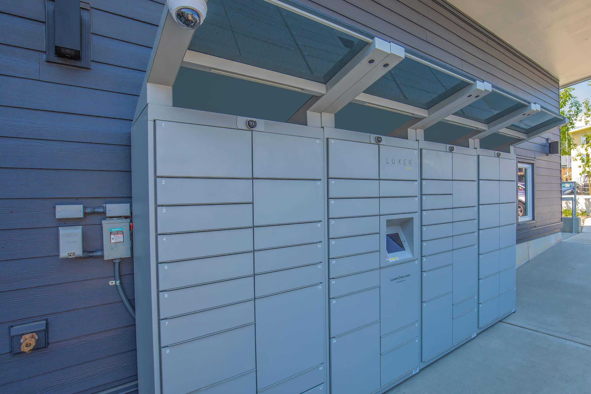 Row of gray package delivery lockers installed against a wall, featuring a digital screen and surveillance camera. The lockers are designed for secure pick-up of parcels and packages. Bright outdoor lighting reflects off the surface, indicating a well-maintained environment.