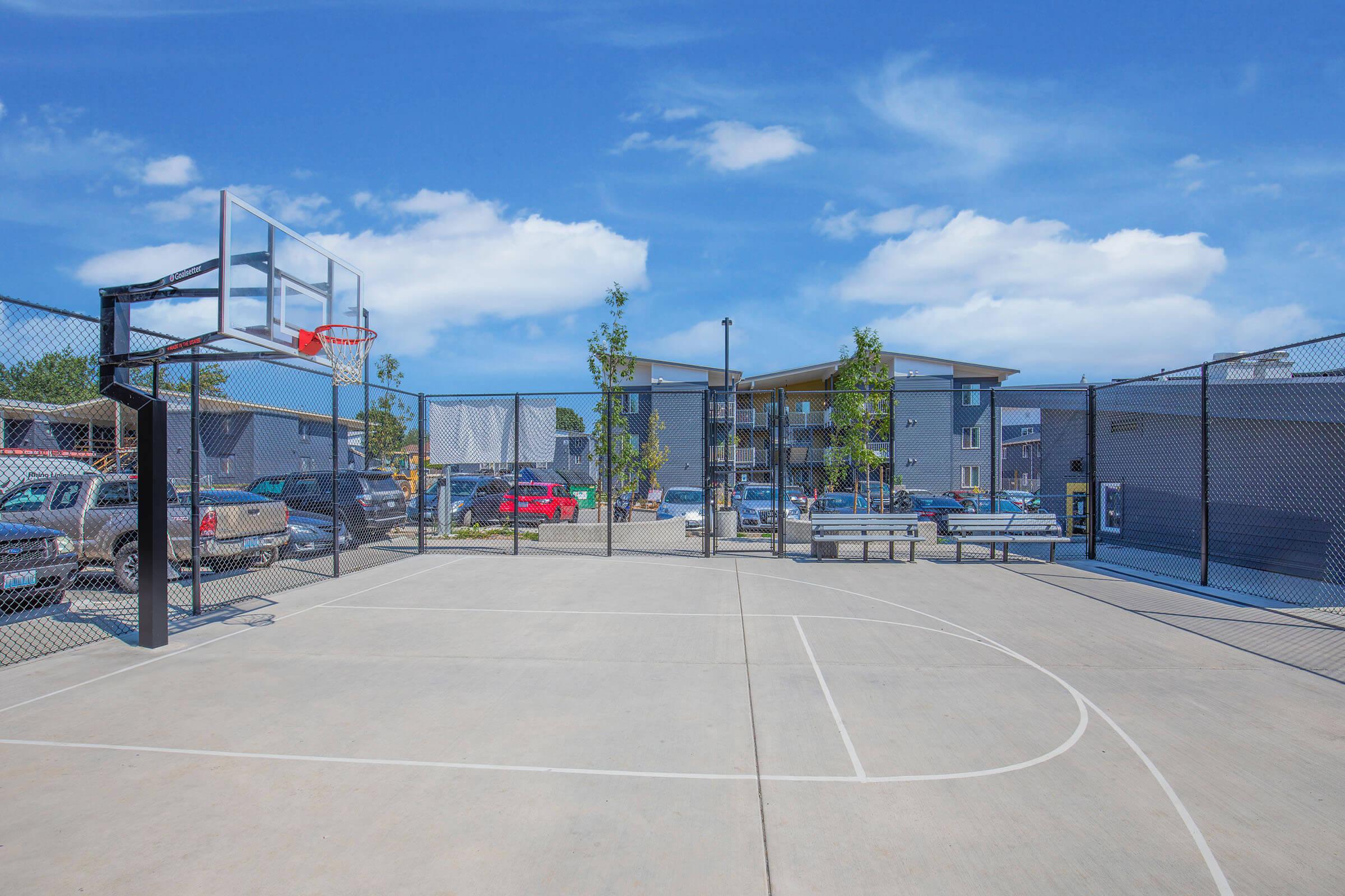 A basketball court with a hoop and backboard, surrounded by a black chain-link fence. In the background, there are several parked cars and modern apartment buildings under a clear blue sky with a few clouds. The court is well-lit and features a smooth concrete surface.