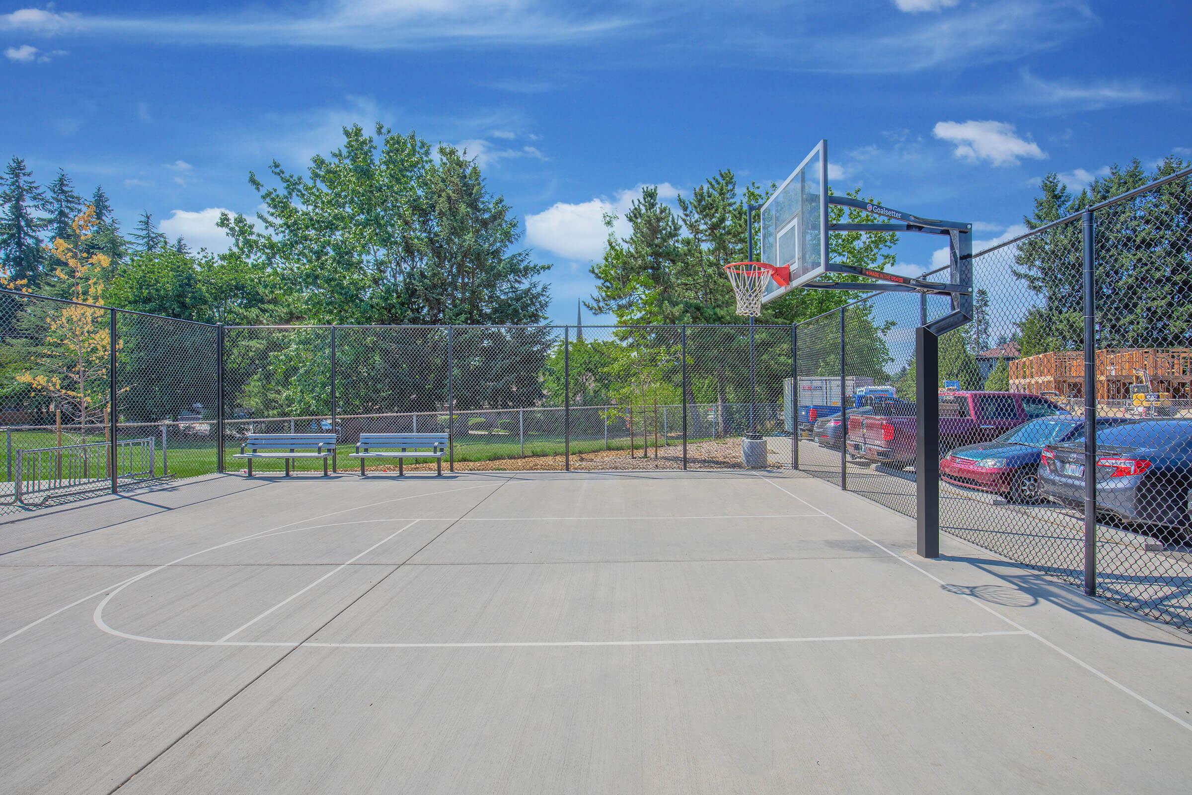 A well-lit outdoor basketball court surrounded by a chain-link fence, featuring a basketball hoop. In the background, there are green trees and parked cars. The court's surface is paved concrete, and a bench is visible nearby. The sky is blue with a few clouds, indicating a sunny day.