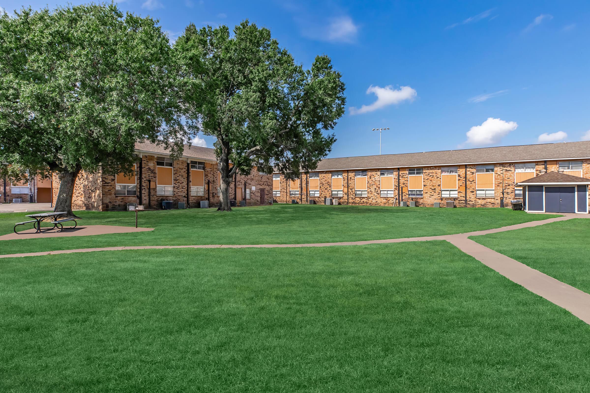 A grassy area features a winding path leading through a park-like setting, surrounded by brick buildings. Large trees provide shade, and a picnic table is visible in the foreground. The sky is partly cloudy, with blue skies enhancing the tranquil atmosphere.