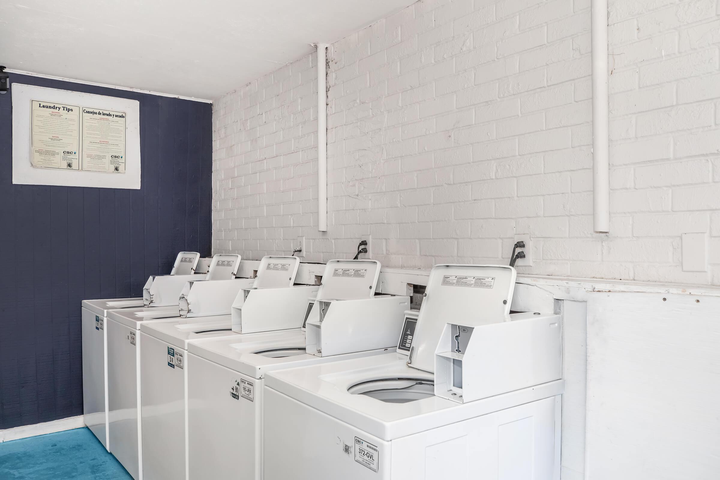 A row of white washing machines lined up against a white brick wall in a laundry room. The floor is a bright blue, and there's a poster hanging on the wall above the machines. The setting appears clean and organized, ideal for laundry tasks.