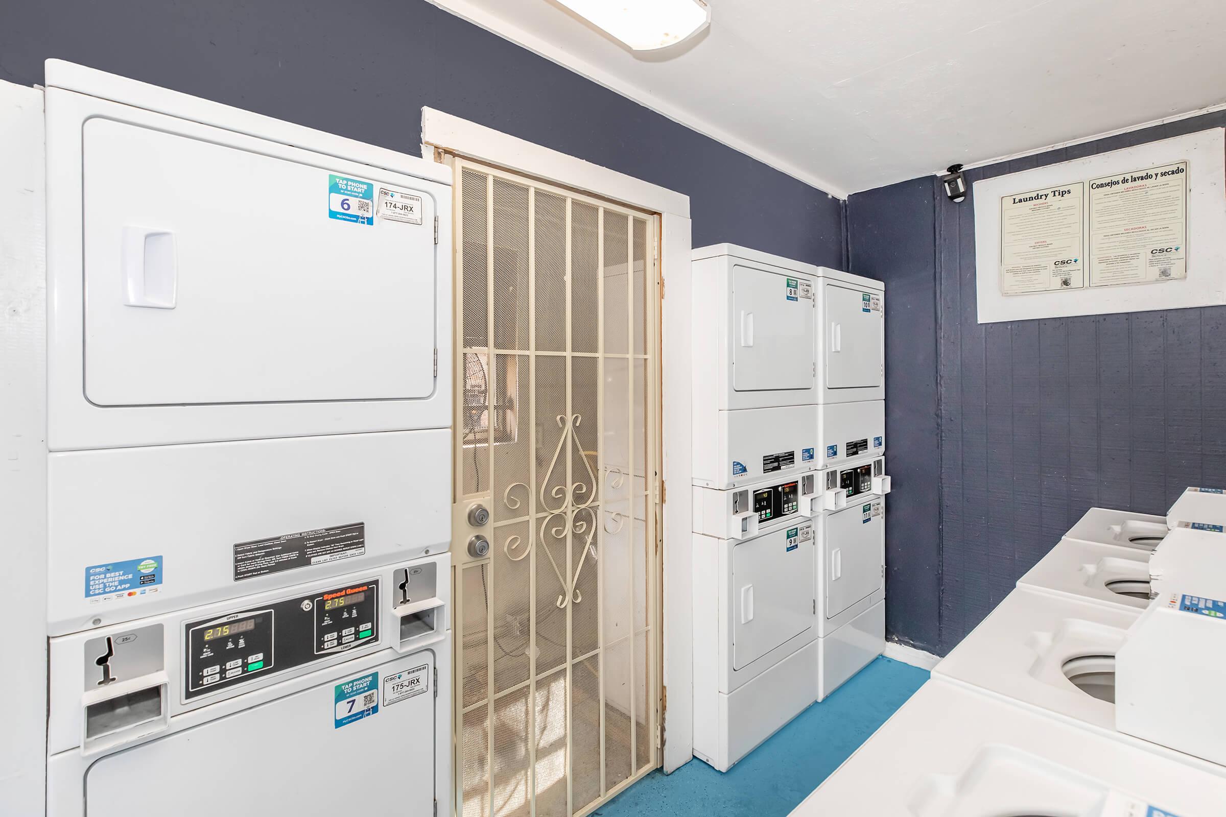 A clean laundry room featuring several stacked washing machines and dryers against a blue and gray wall. A door with a metal security gate is visible on the left. Important laundry instructions are posted on the wall, and the floor is covered with a blue mat, creating a functional and organized space for laundry.
