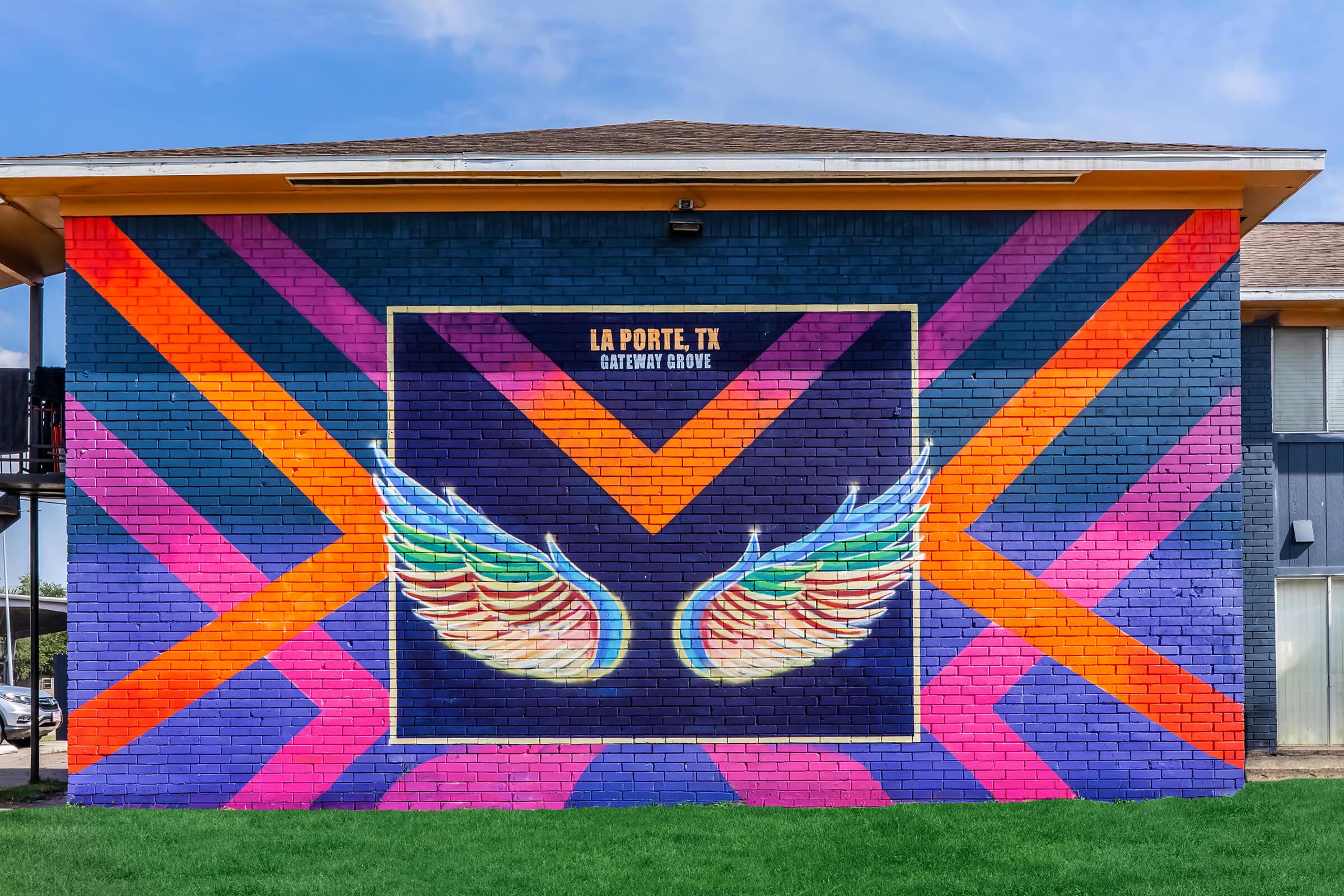 A colorful mural on a brick wall featuring a pair of angel wings at the center, surrounded by vibrant geometric patterns in shades of pink, orange, purple, and blue. The mural is located in La Porte, TX, and adds artistic flair to the building's exterior.