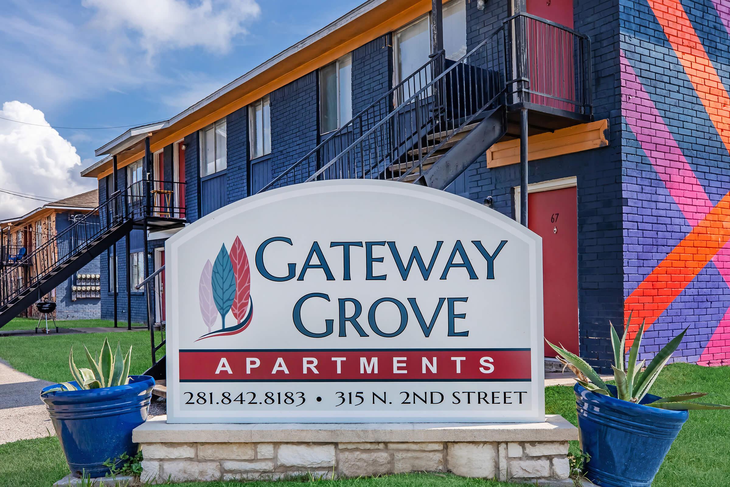 Sign for Gateway Grove Apartments located at 315 N. 2nd Street, featuring a decorative logo with leaves. The building behind the sign has a colorful mural on the side, with well-maintained landscaping and potted plants.