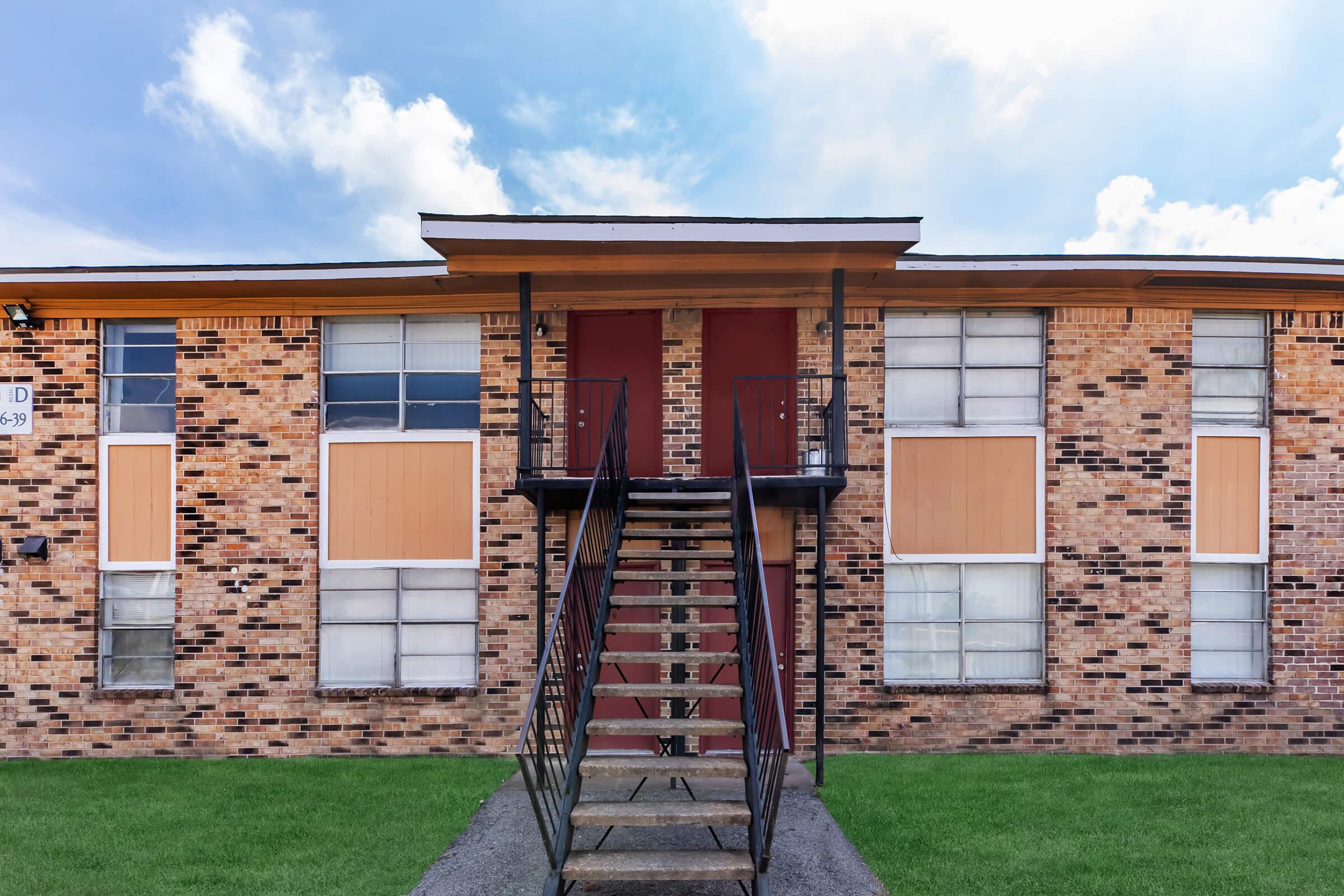 A two-story brick apartment building with a central staircase leading to two red doors. Some windows are boarded up, with a grassy area in front. The sky is blue with a few clouds.