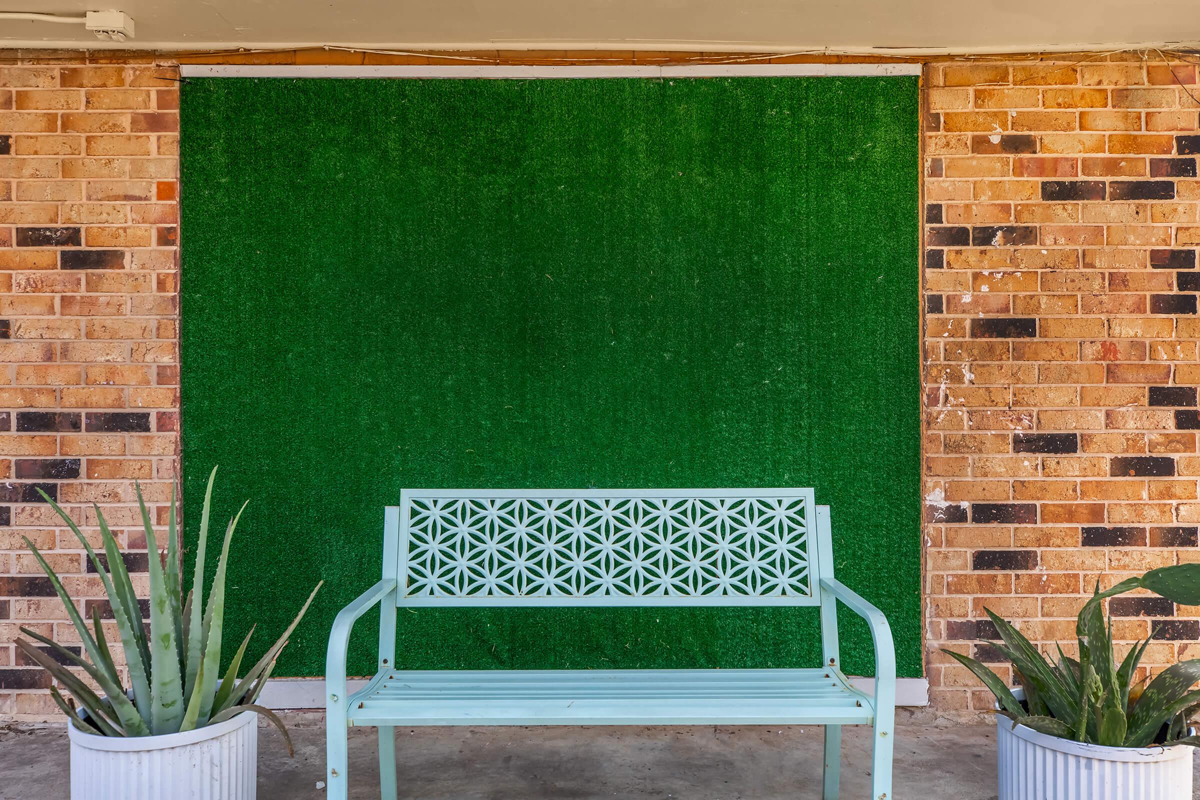 A mint green bench is positioned in front of a textured green wall, made to look like grass. There are two large potted plants on either side of the bench, and the background features a mix of light and dark brown brick patterns. The scene is simple and inviting, ideal for relaxation.