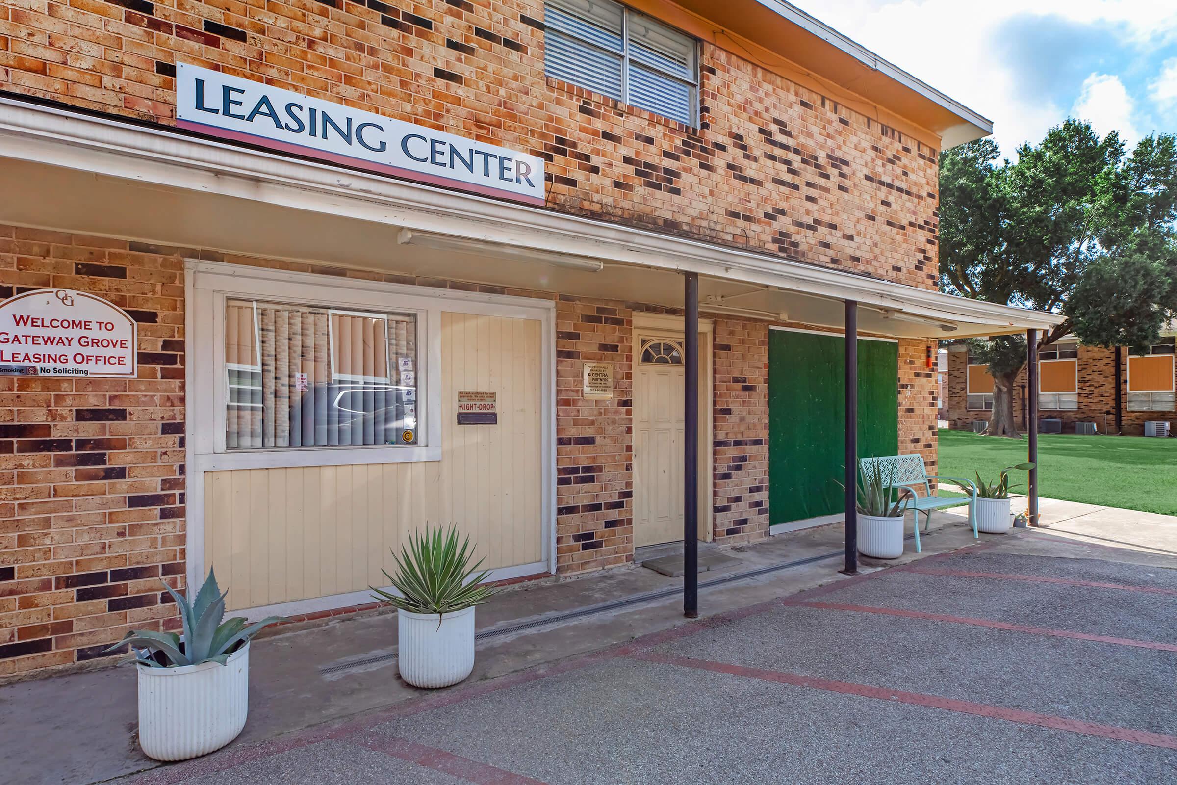 A brick building featuring a Leasing Center sign. The entrance has a white door with a small window, flanked by potted plants. A sign on the left reads "Welcome to Gateway Grove Leasing Office." The area includes a grassy patch and additional buildings in the background under a blue sky.