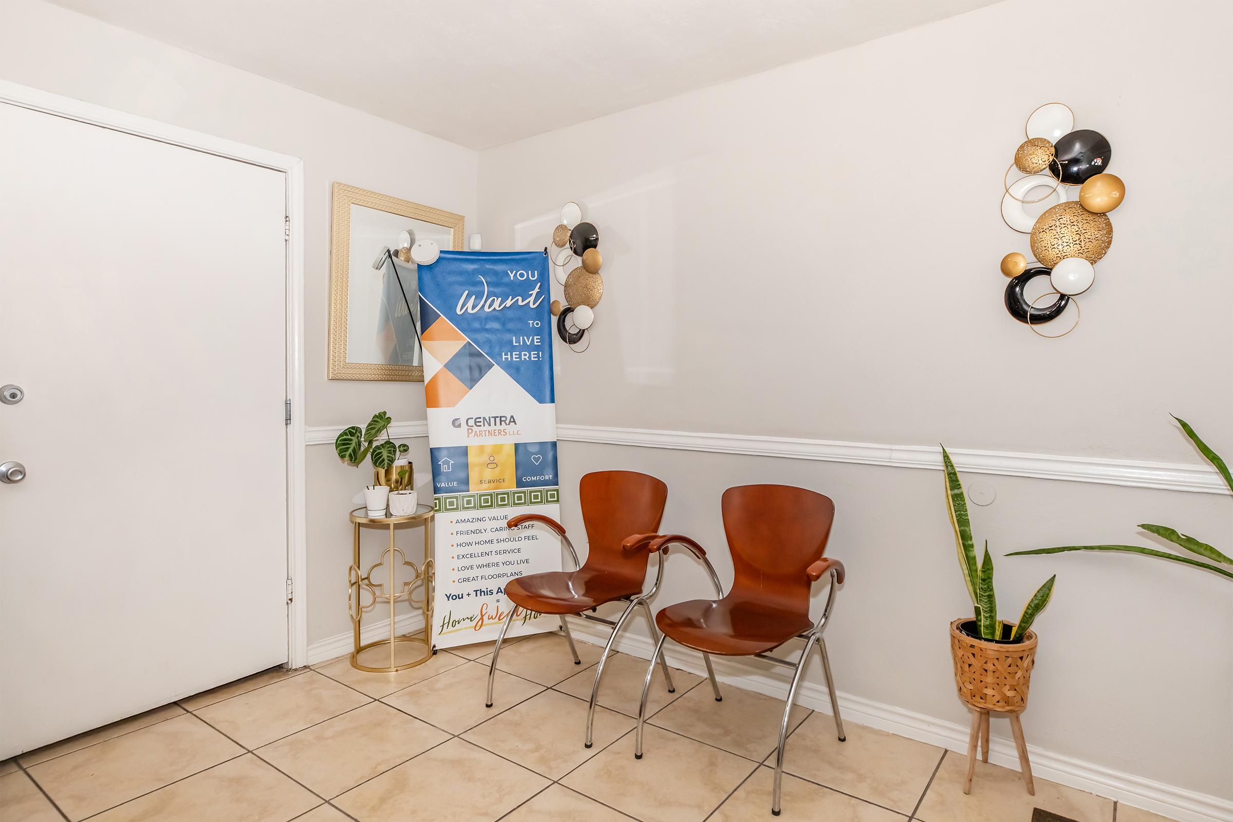 A welcoming entryway featuring a door, a mirror, two brown chairs, and a decorative plant. A promotional banner is displayed prominently, encouraging potential residents to inquire about living options. Wall decor includes circular elements in black, gold, and white. The tiled floor completes the inviting atmosphere.