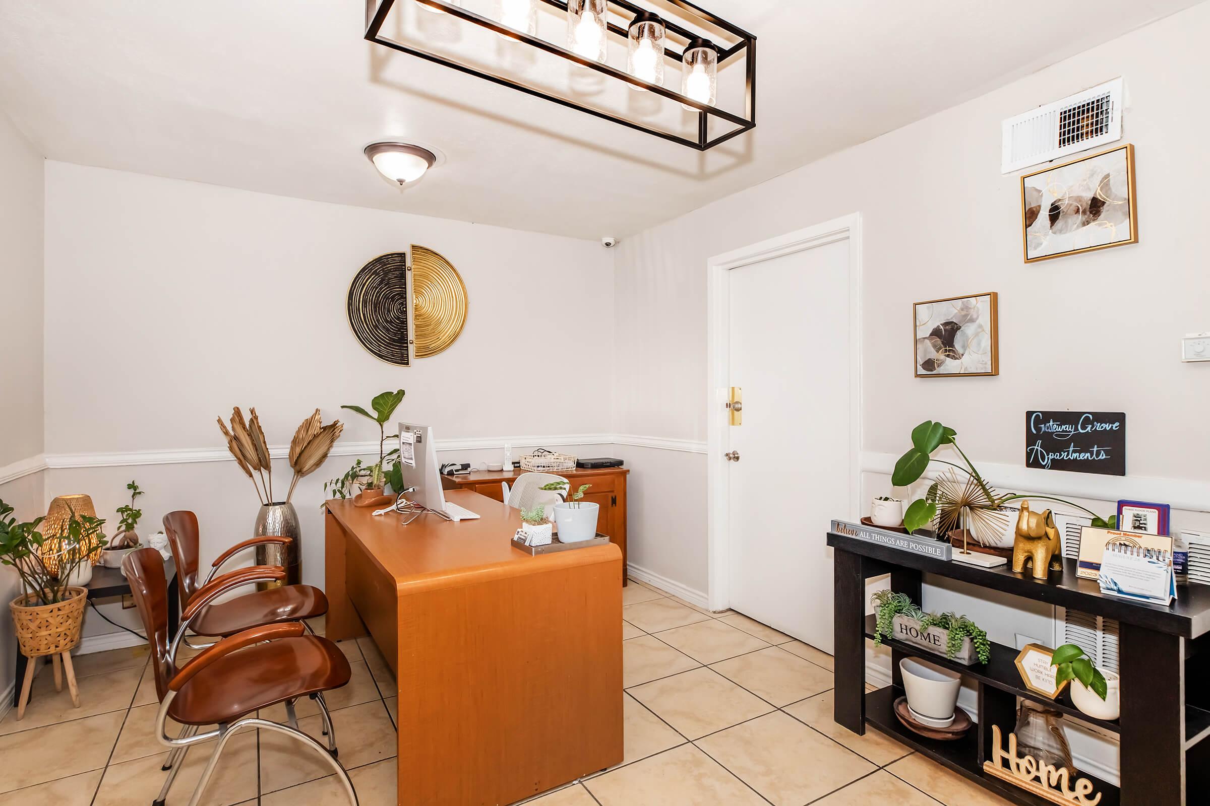 A cozy, inviting office space featuring wooden furniture, two stylish chairs, and a sleek desk with a computer. The walls are adorned with framed art and there are decorative plants throughout. A shelf displays various decorative items, adding a warm touch to the professional atmosphere.