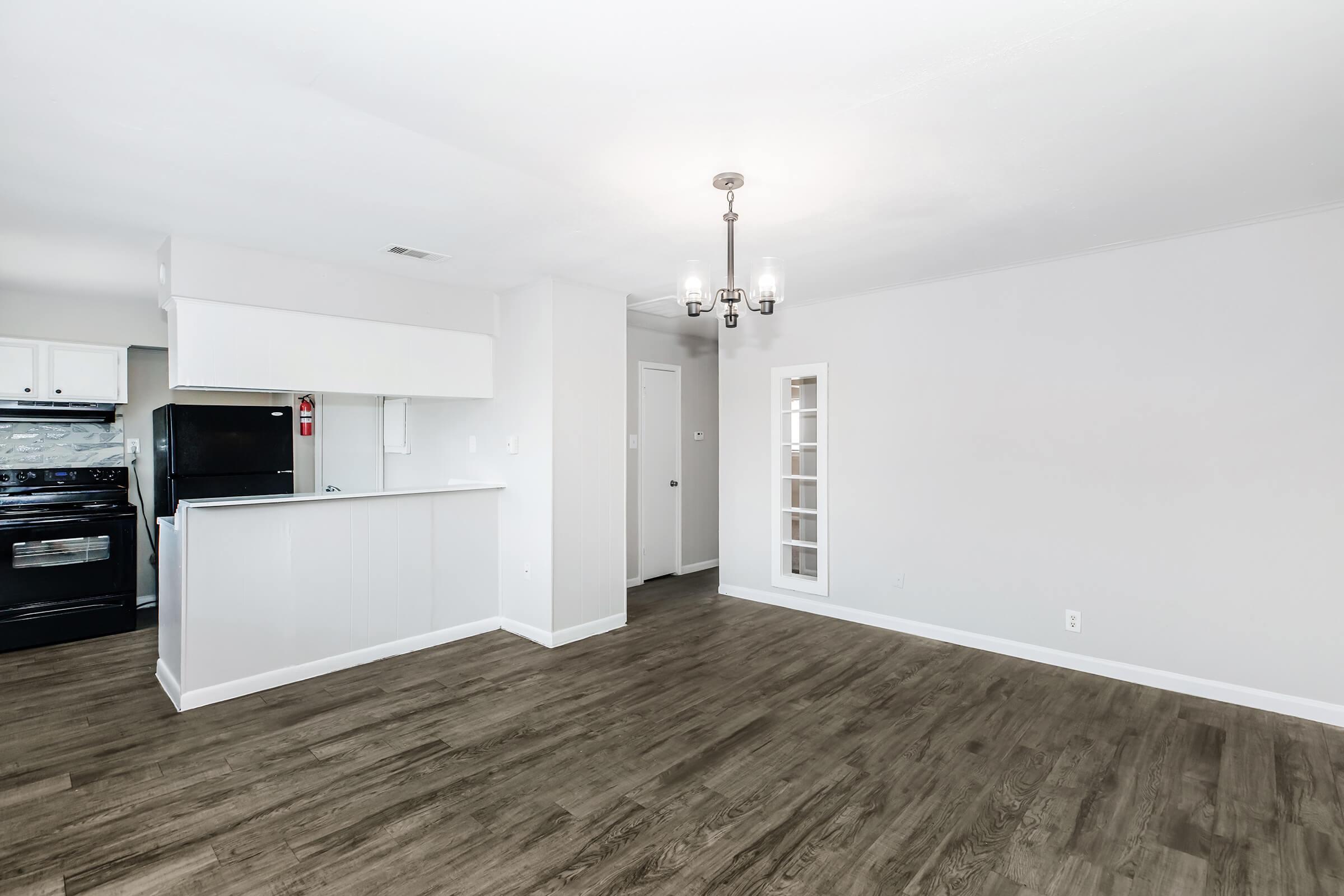 A modern, empty living space featuring light-colored walls and dark wood flooring. The room includes a dining area with a chandelier, a kitchen nook with a black stove and refrigerator, and a door leading to another area. A small, glass-paneled cabinet is visible on the right side of the room.