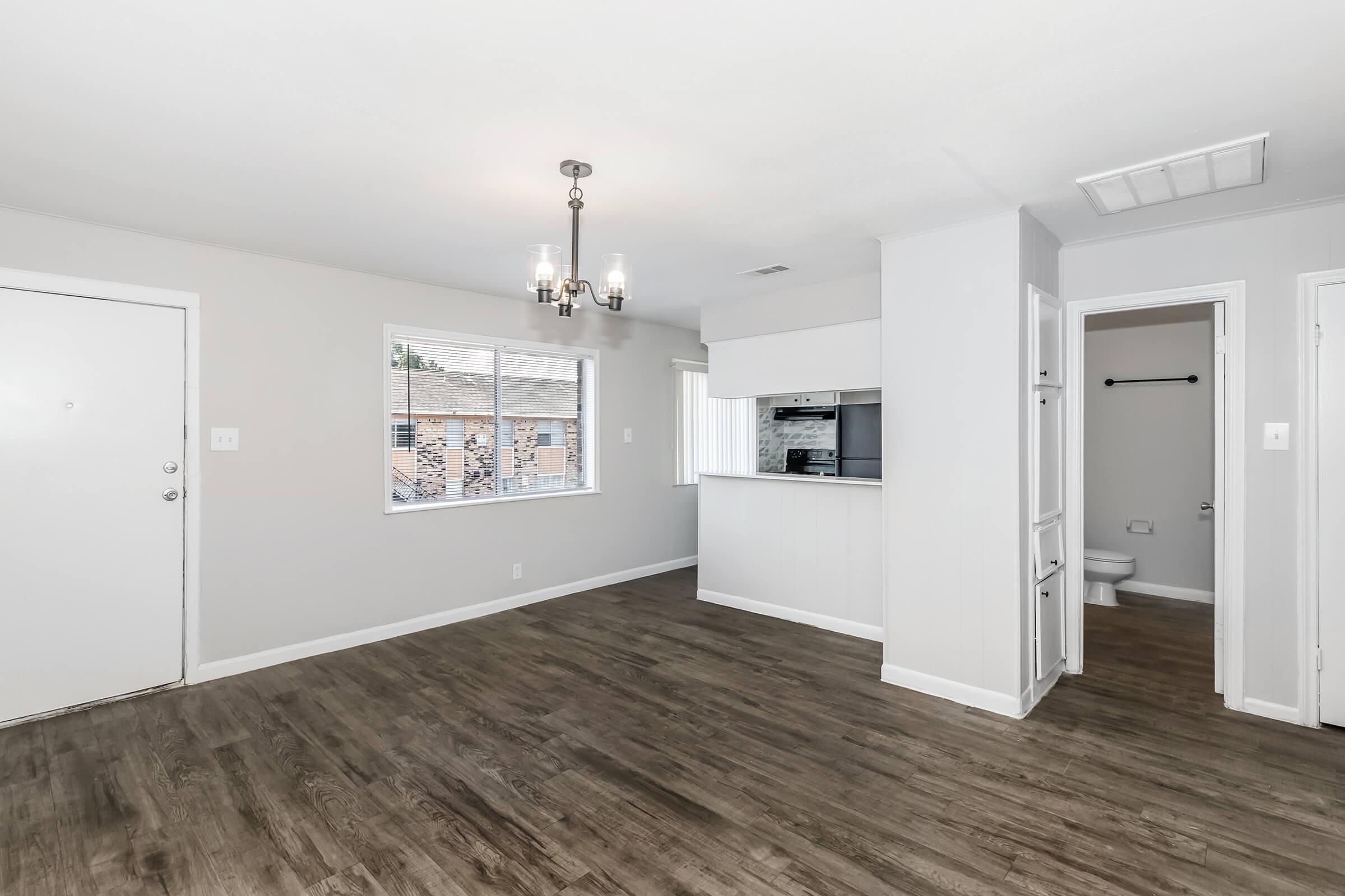 A spacious, well-lit living area with light gray walls, hardwood-style flooring, and a modern chandelier. A large window provides natural light, while a doorway leads to a bathroom. The kitchen is partially visible in the background, featuring a bar counter. The entrance door is on the left.