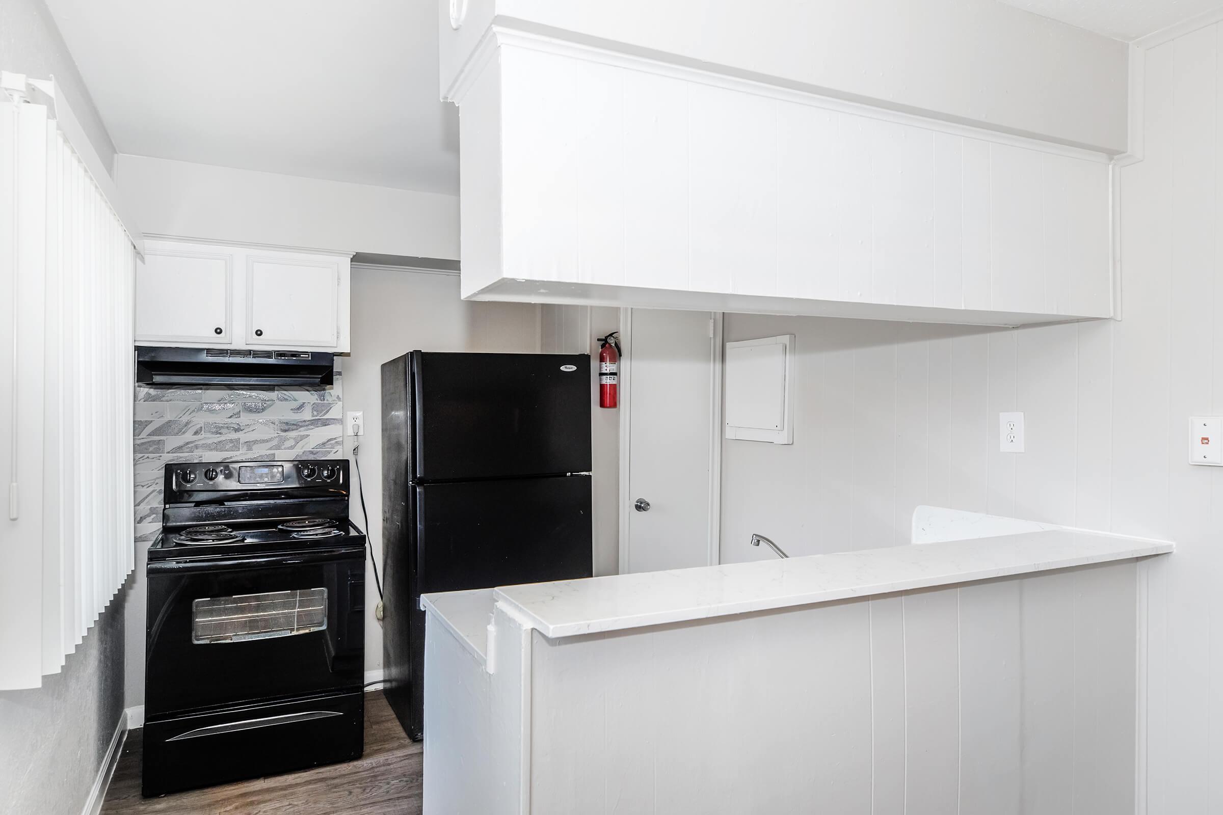 A modern kitchen featuring a black refrigerator and stove, white cabinetry, and a marble backsplash. There is a small countertop area separating the kitchen from another room, with light-colored walls and a window covered by white blinds. A fire extinguisher is mounted on the wall.