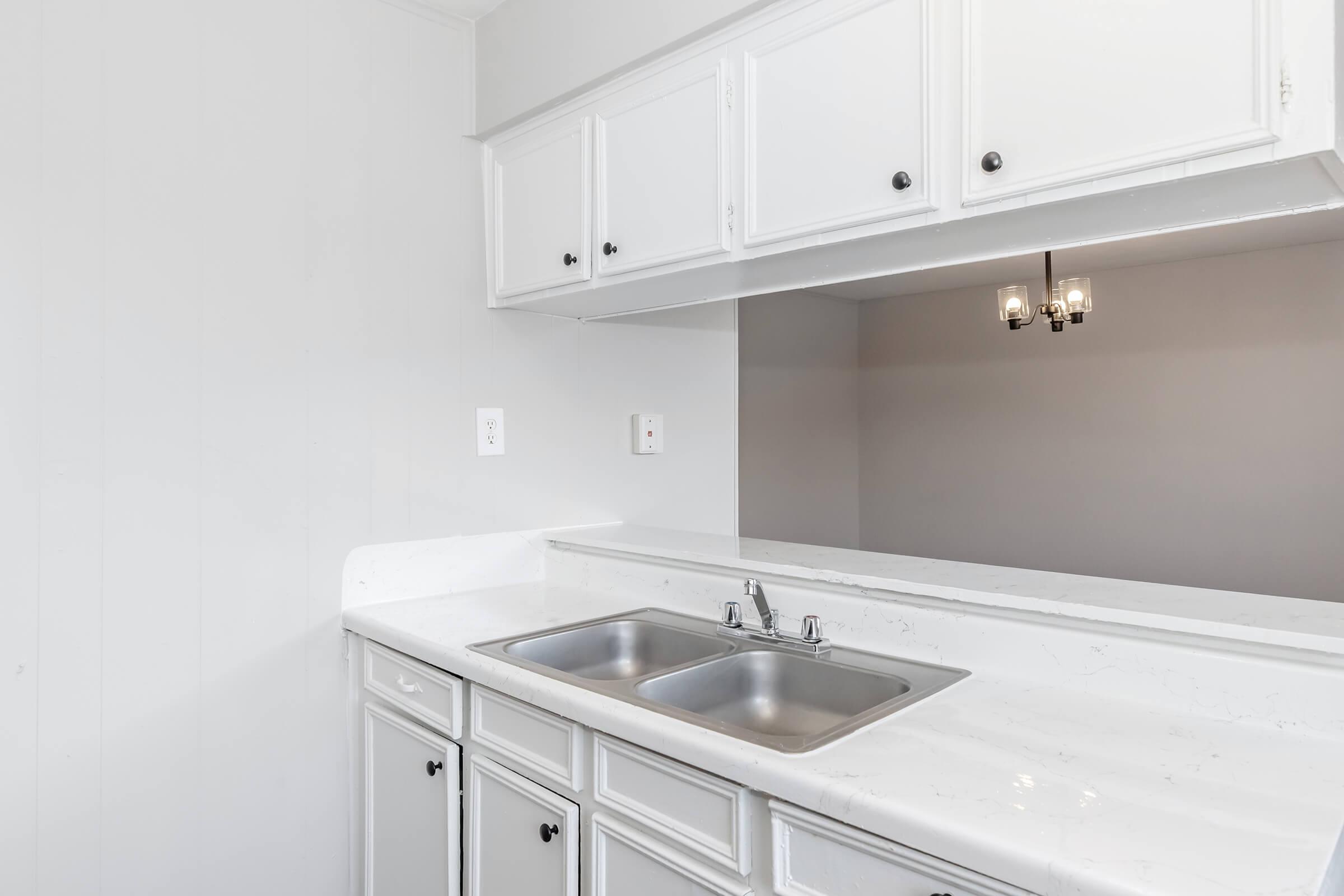 A clean, modern kitchen with white cabinets and a marble countertop featuring a double sink. The walls are light gray, and there is a small chandelier visible above. The overall space appears bright and minimalistic, creating a fresh atmosphere.