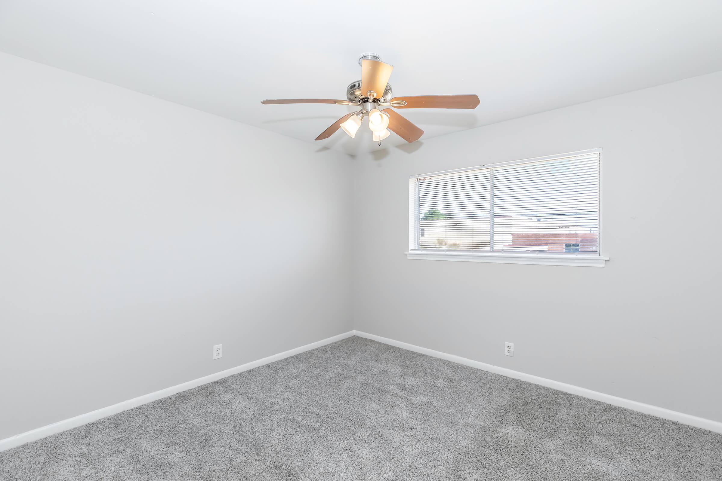 A vacant room with light gray walls and plush gray carpeting. The ceiling features a ceiling fan with wooden blades, and there is a window fitted with white blinds that allows natural light to enter. The room appears spacious and modern, ready for furnishing.