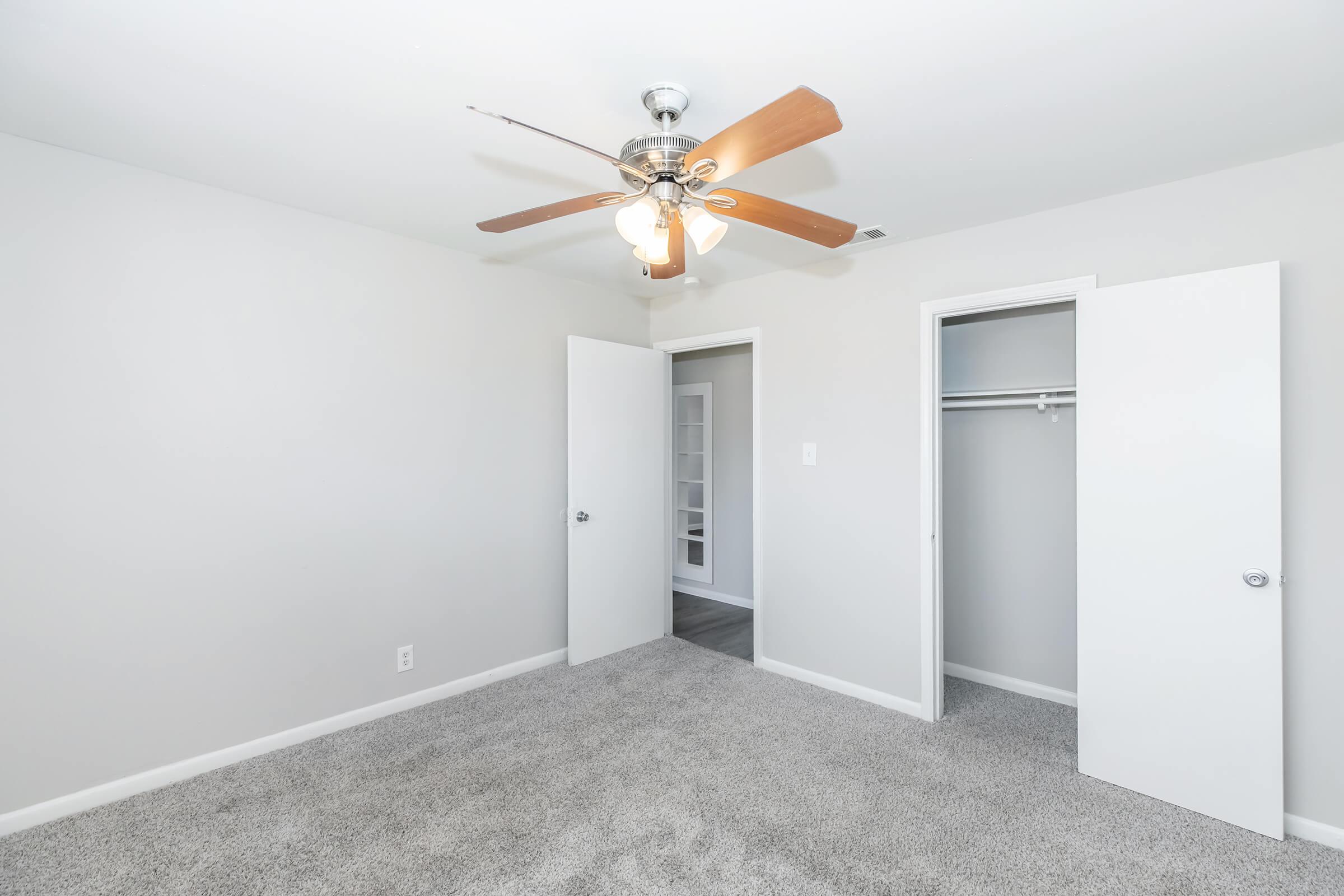 A well-lit room with light gray walls and carpet. A ceiling fan with wooden blades hangs from the ceiling. Two white doors are open, revealing a closet space and another room beyond. The space is empty, offering a neutral backdrop suitable for various uses.