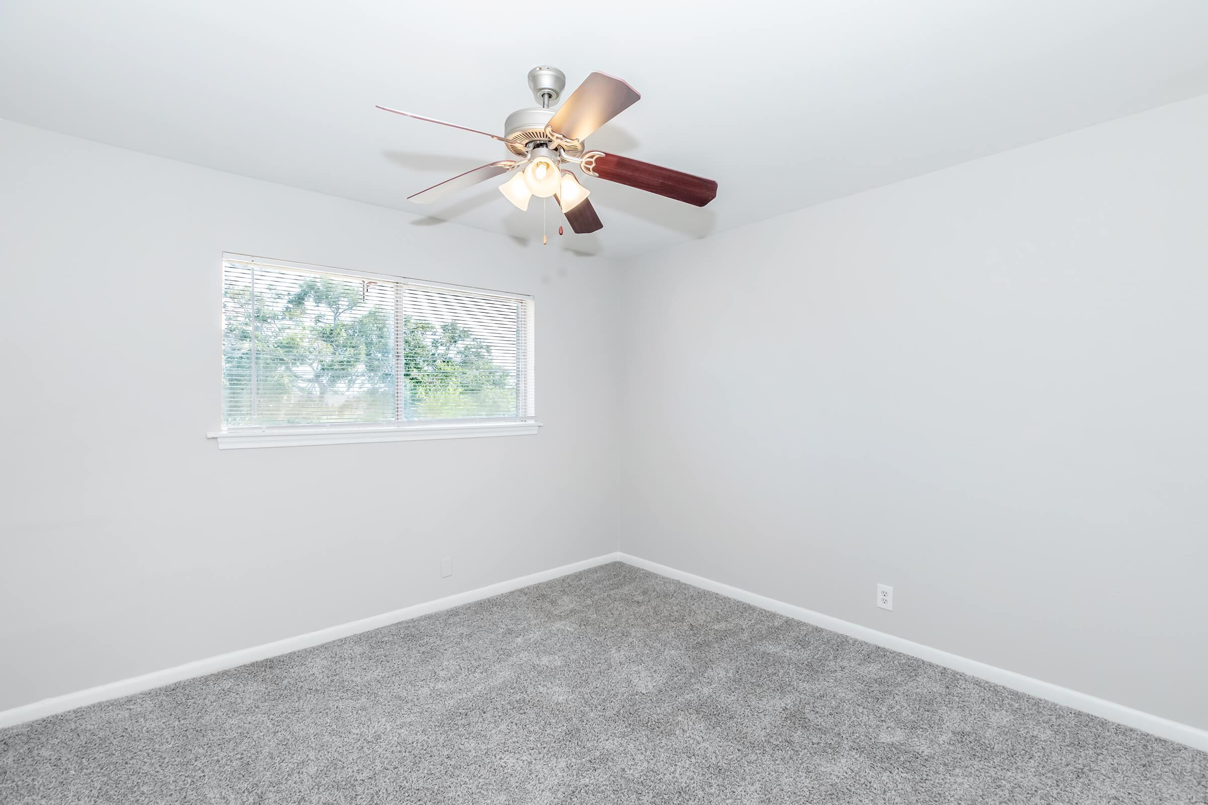 A spacious, minimalist bedroom featuring light gray walls and carpet. A ceiling fan with wooden blades hangs from the ceiling, and a window allows natural light to enter, framed by white trim. The room is devoid of furniture, creating an open and airy atmosphere.