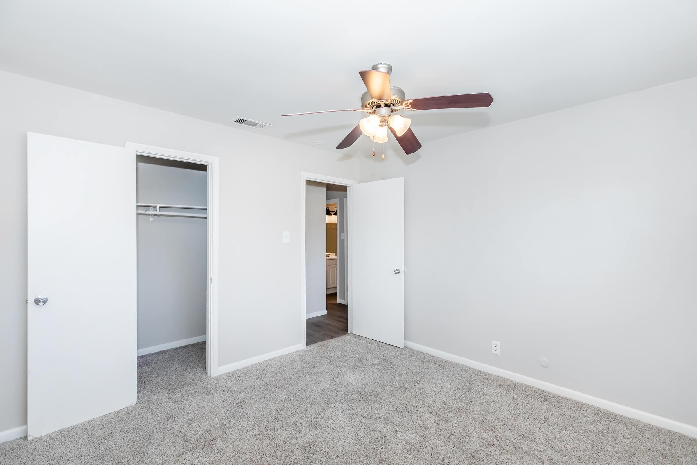 A bright, empty bedroom with light gray walls and plush carpet. Two white doors lead to closets, and a ceiling fan with three light bulbs is mounted above. The room appears spacious and well-lit, enhancing its inviting atmosphere.