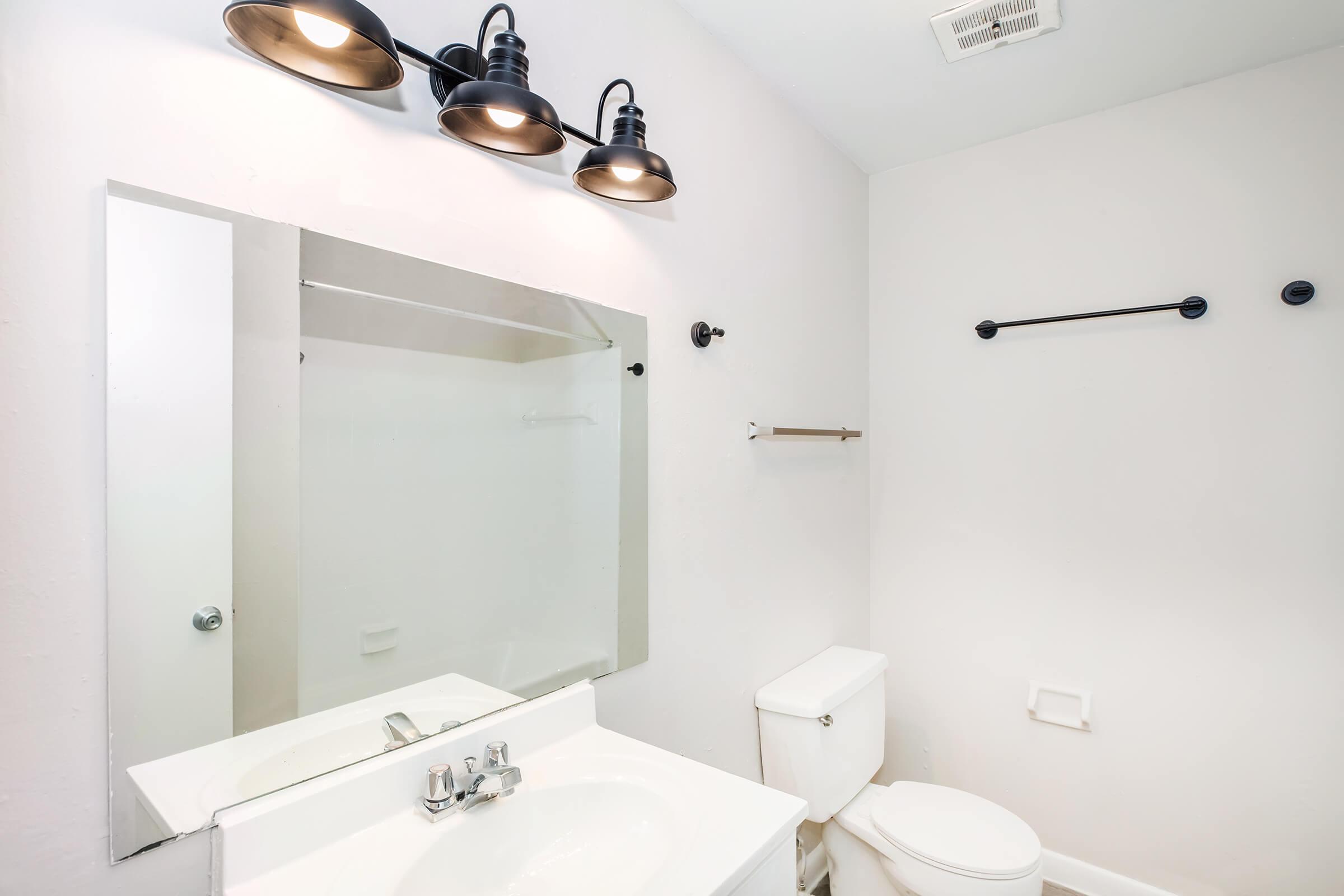 A modern bathroom featuring a white sink with a chrome faucet, a large mirror above the sink, a white toilet, and a shower with a glass door. Three black light fixtures illuminate the space, and a towel bar is mounted on the wall. The walls are painted a light color, creating a bright and airy atmosphere.