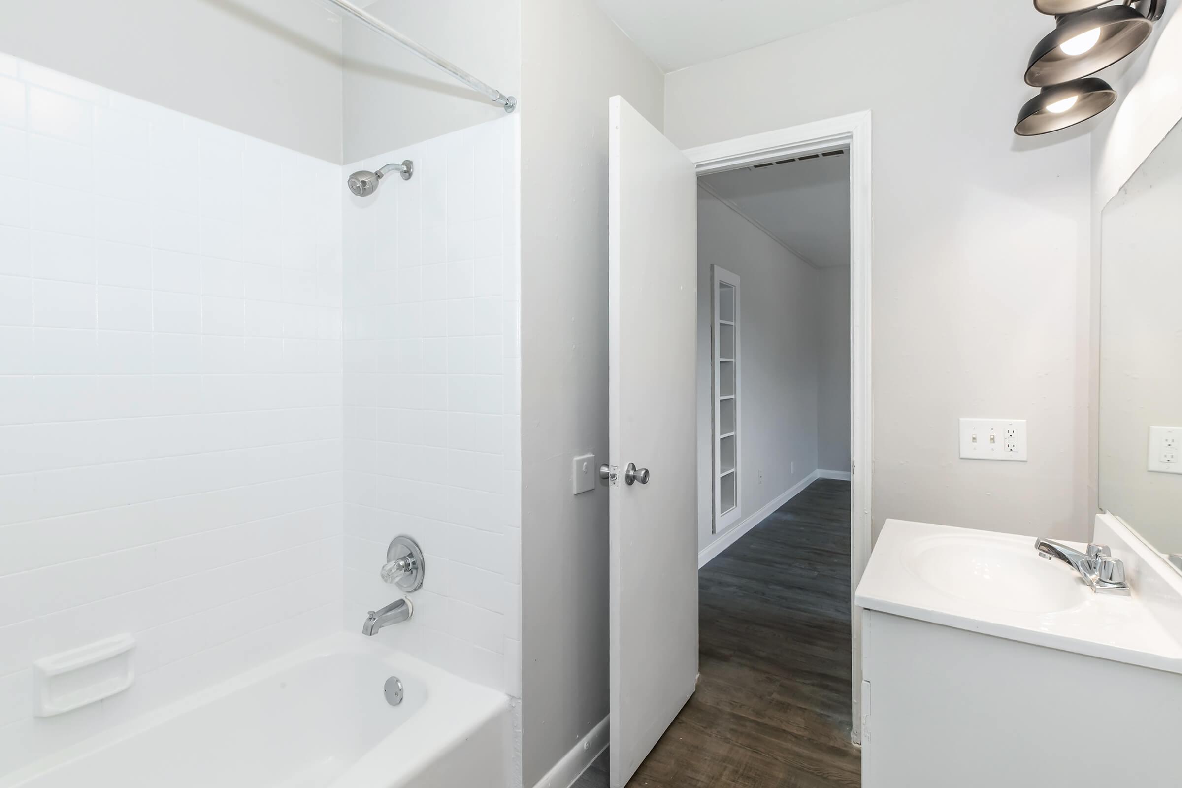 A clean bathroom featuring a white tub and shower with a glass shower rod. A modern sink and mirror are on the right side, positioned against a light-colored wall. A door leads to a hallway, showing a glimpse of a space beyond with light flooring and additional doors.