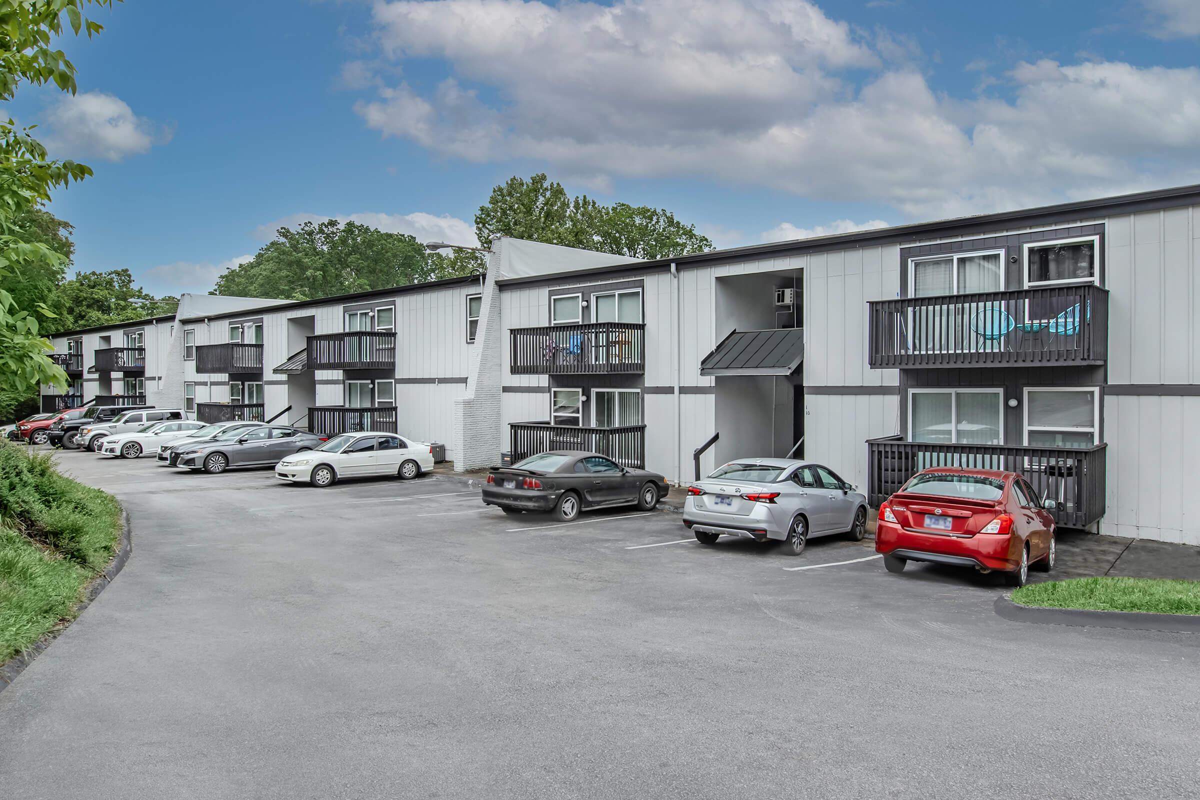 A residential apartment complex with two-story buildings. The parking lot is filled with several cars, and some units have balconies. Lush greenery surrounds the area, and the sky is partly cloudy, creating a peaceful suburban atmosphere.