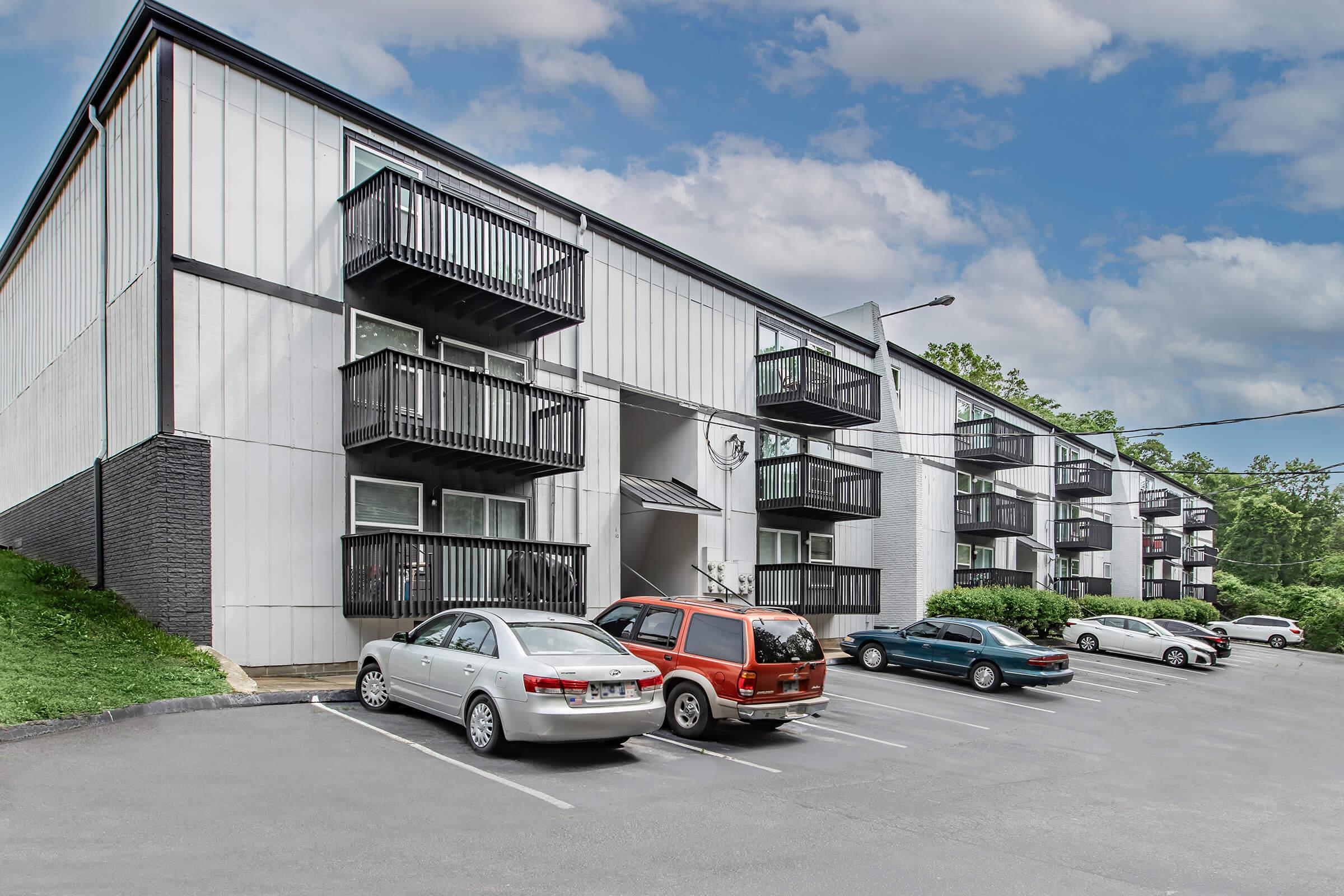 A modern multi-story apartment building with balconies, surrounded by greenery. Several cars are parked in front, and the scene is set under a blue sky with scattered clouds. The building features a mix of light and dark siding, creating a contemporary appearance.