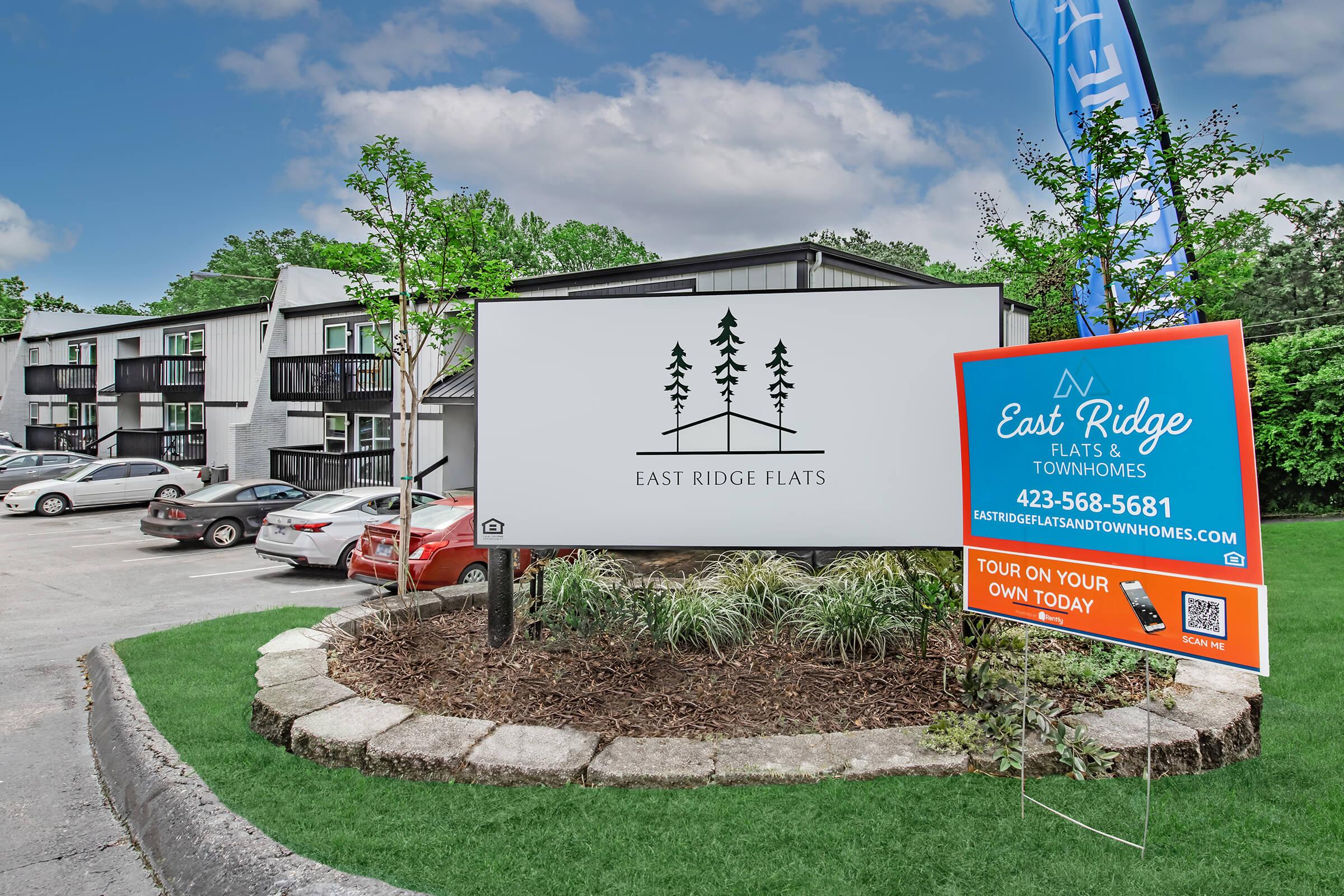 Signage for East Ridge Flats and Townhomes, featuring a logo with trees, along with a promotional banner. The setting includes landscaped grass and a parking area with cars. Contact information and a QR code are displayed on the sign encouraging tours.