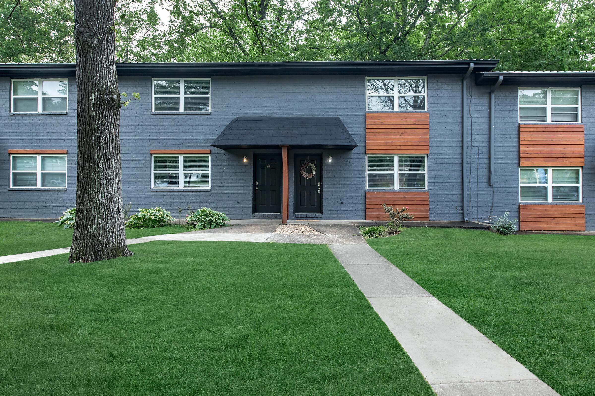A modern apartment building with a gray exterior, wooden accents, and several windows. A well-maintained lawn surrounds the building, with a paved pathway leading to the entrance. Trees provide shade in the background, creating a serene and inviting atmosphere.