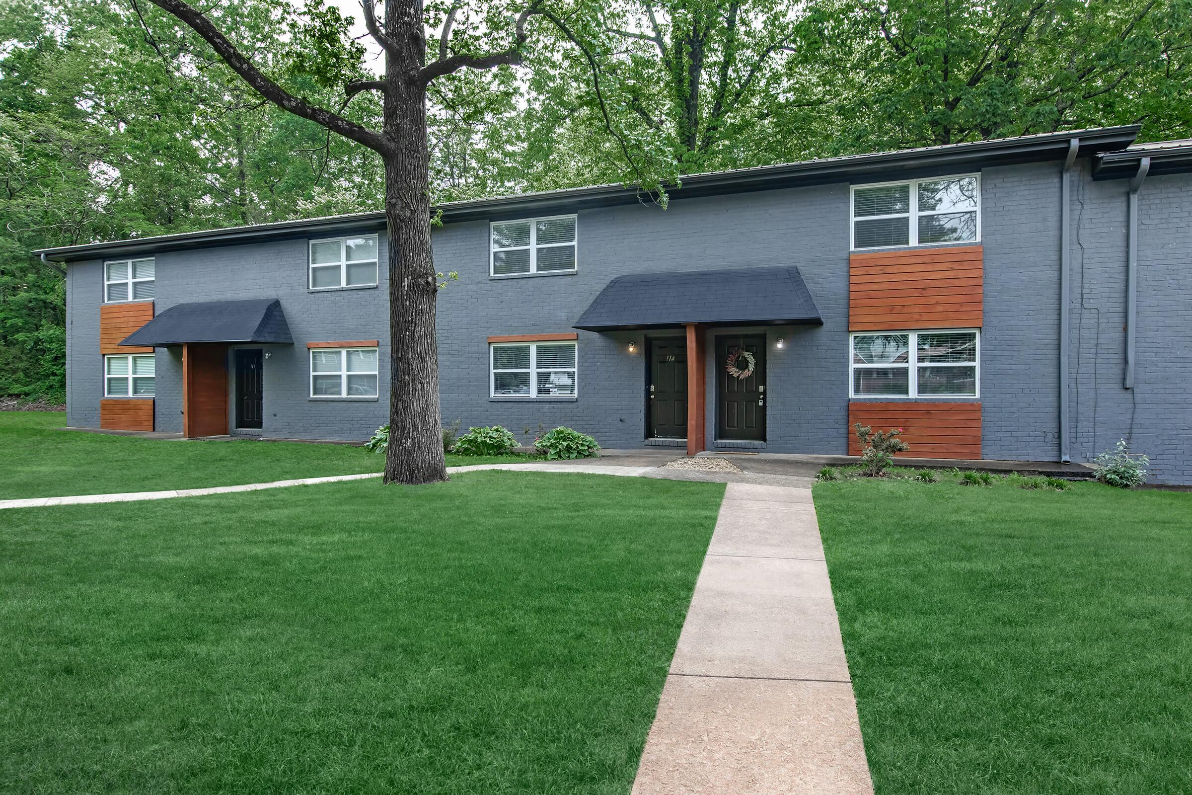A modern apartment building with a gray exterior and wooden accents. The building features a symmetrical design with two entrances, surrounded by a well-maintained lawn and walking path. Trees in the background add to the greenery of the setting.