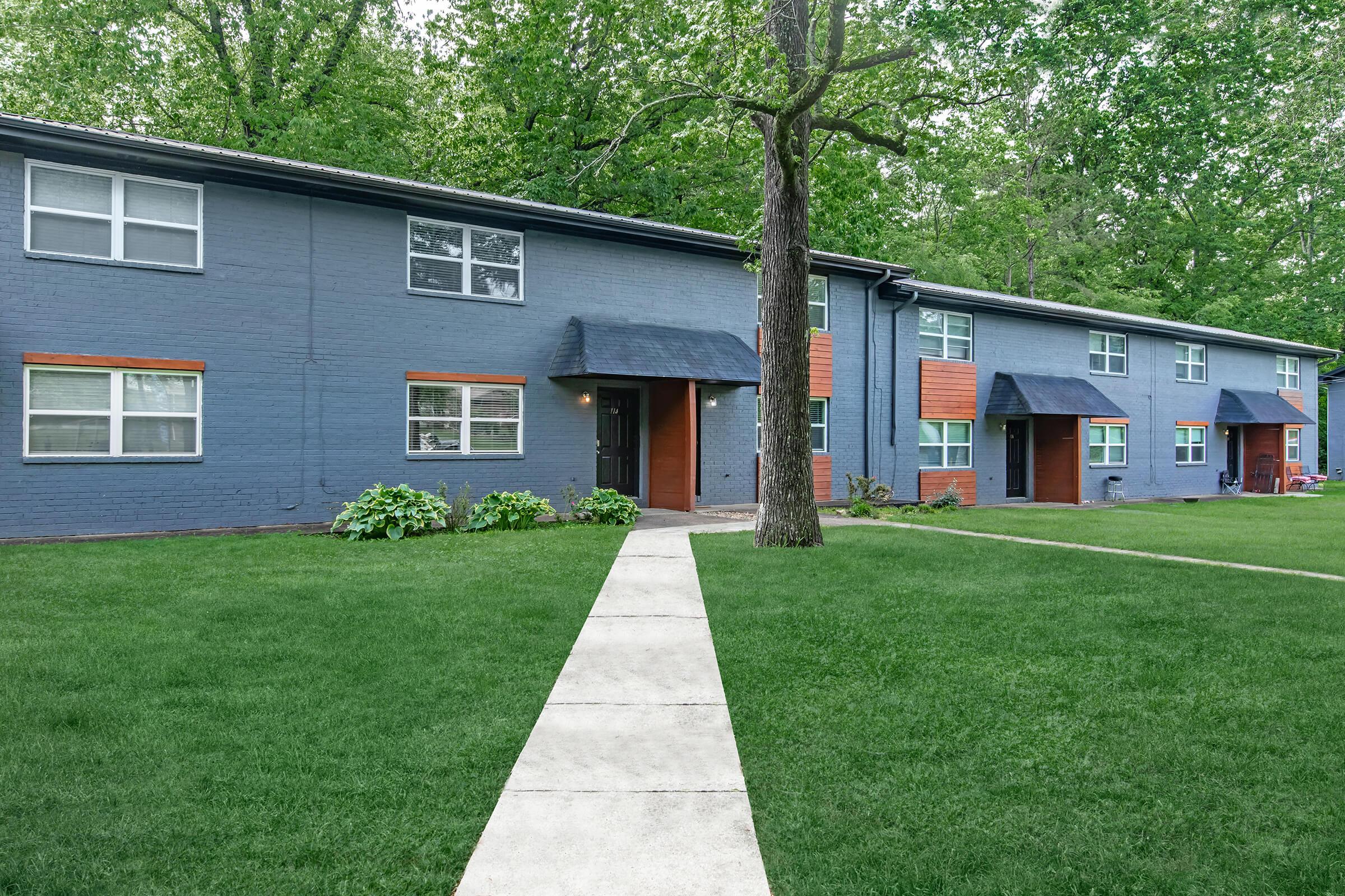 Two-story apartment building with blue-gray exterior and wooden accents, surrounded by green grass and trees. A concrete walkway leads to the entrance, with several doors visible. The setting is lush and residential, conveying a peaceful atmosphere.