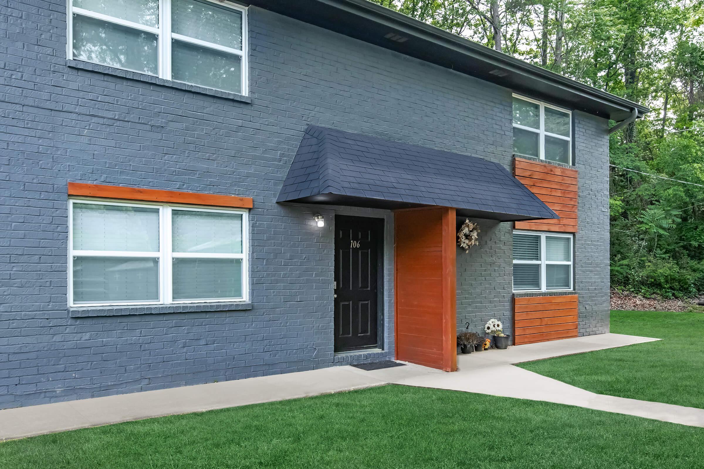 Exterior of a modern two-story building with gray brick walls and wooden accents. The entrance features a black door under a small wooden awning, flanked by two windows. A well-maintained lawn is in front, and trees are visible in the background, creating a natural setting.