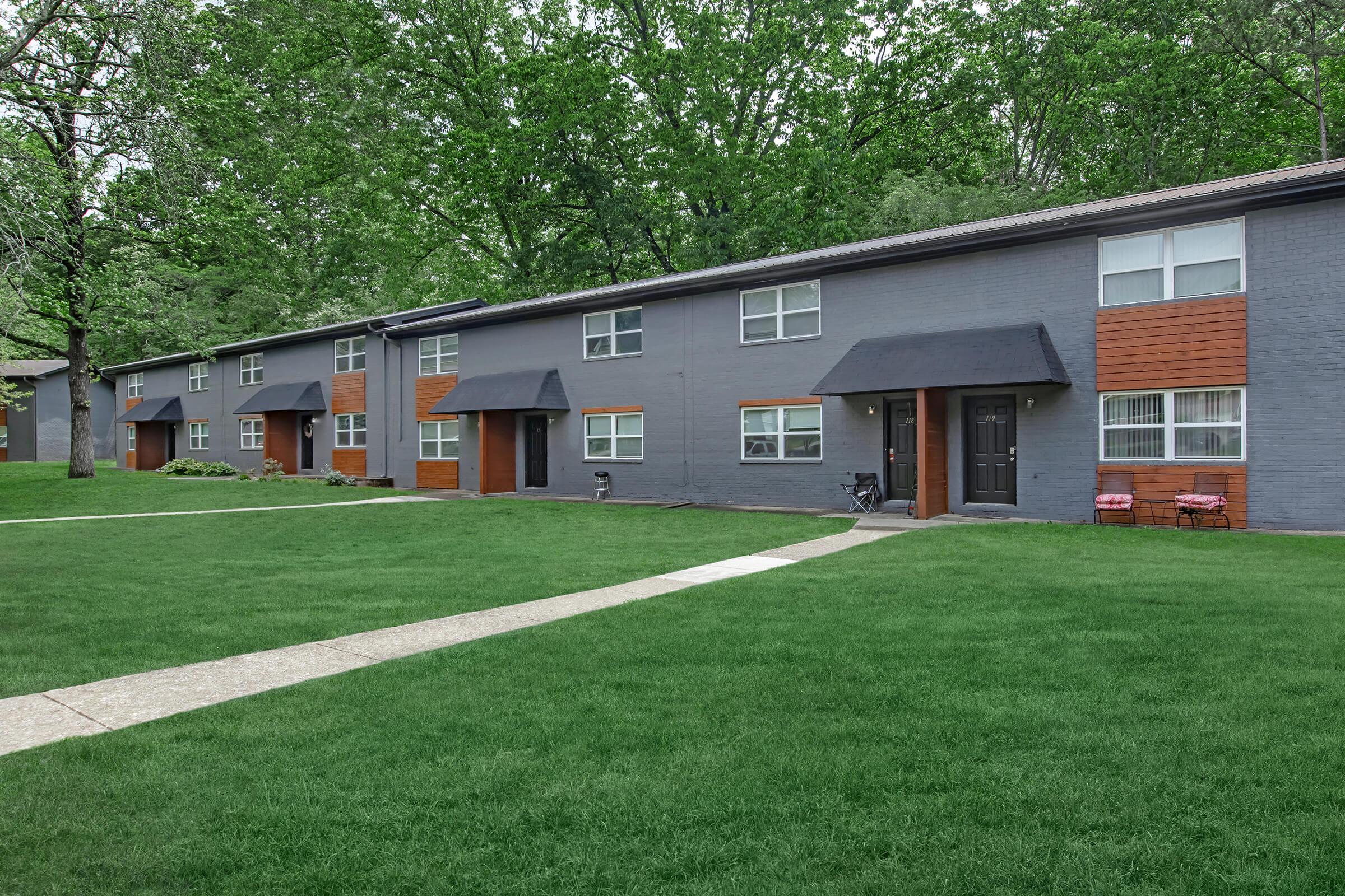 Row of modern, gray residential buildings with wooden accents, set against a backdrop of lush green trees. The well-maintained lawn features a concrete walkway leading to the entrances of the units, which have small black awnings. Two chairs are placed outside one of the units.