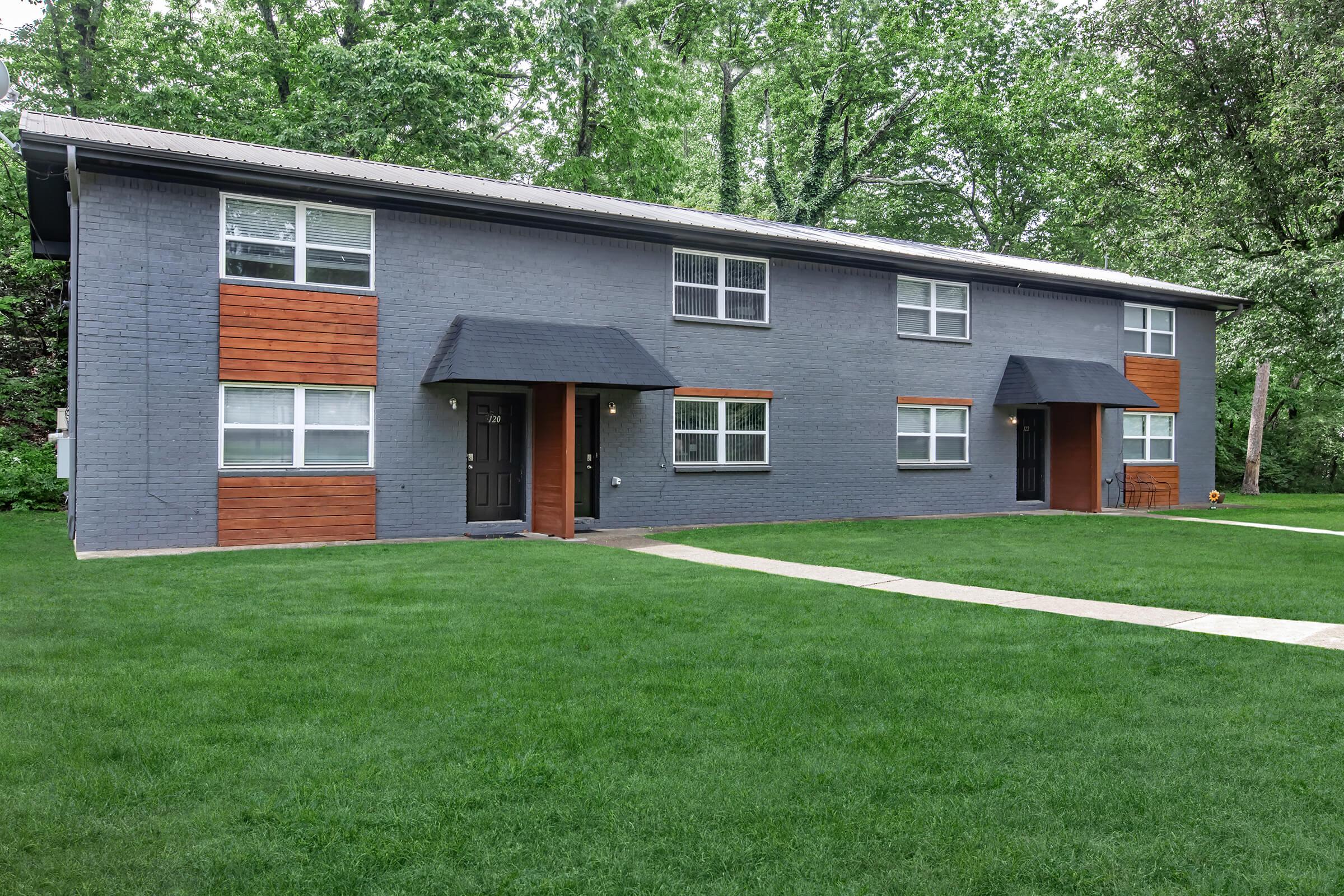 A modern two-story apartment building with a grey exterior, featuring wooden accents on the lower half of its façade. The front has multiple windows with white frames and two main entrances, surrounded by a well-maintained lawn with lush green grass. Trees line the background, adding to the natural setting.