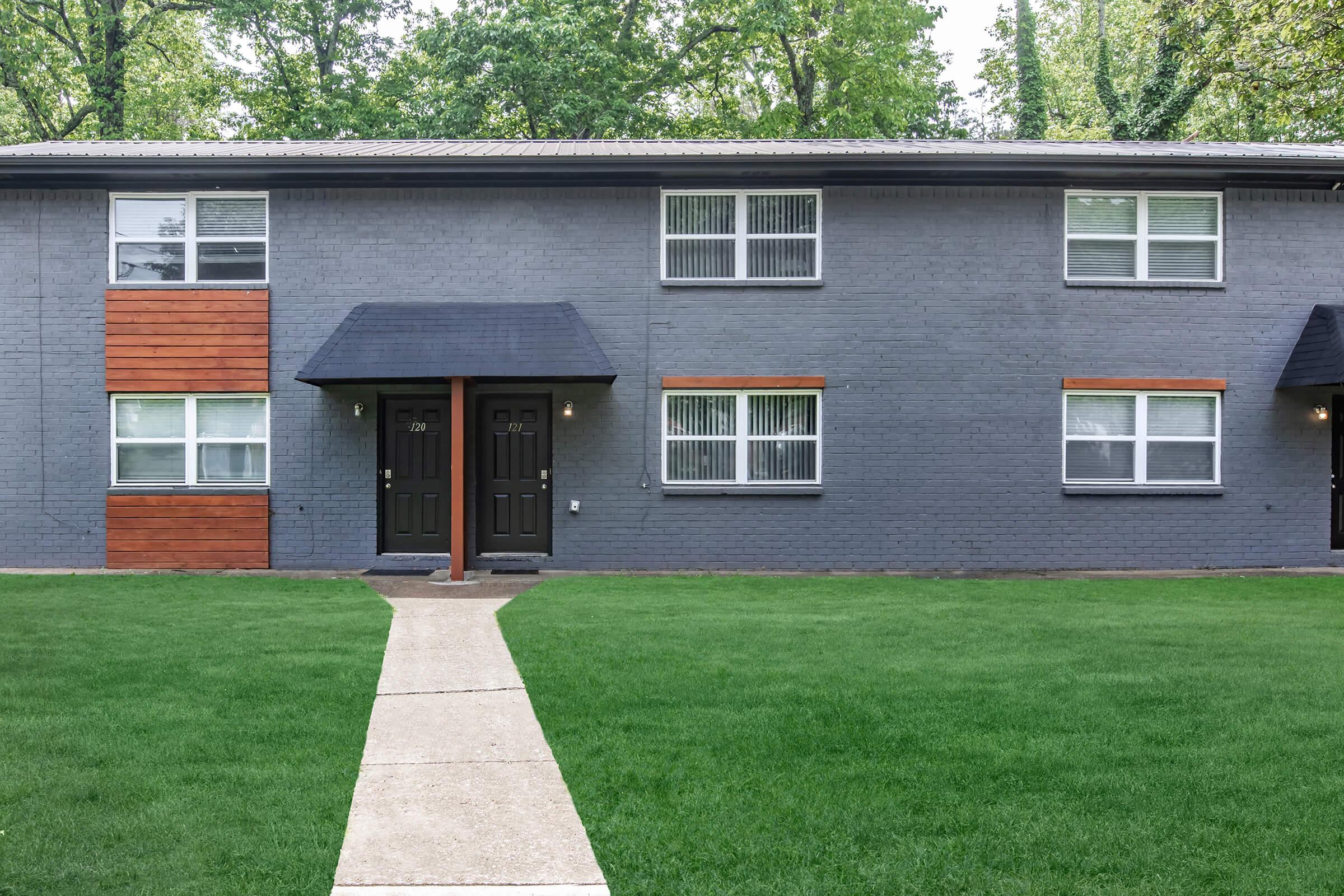 Two-story apartment building with a gray exterior and wooden accents. The entrance features two black doors on the ground floor, flanked by large windows. A concrete pathway leads to the doors, and the area is surrounded by well-maintained green grass and trees in the background.
