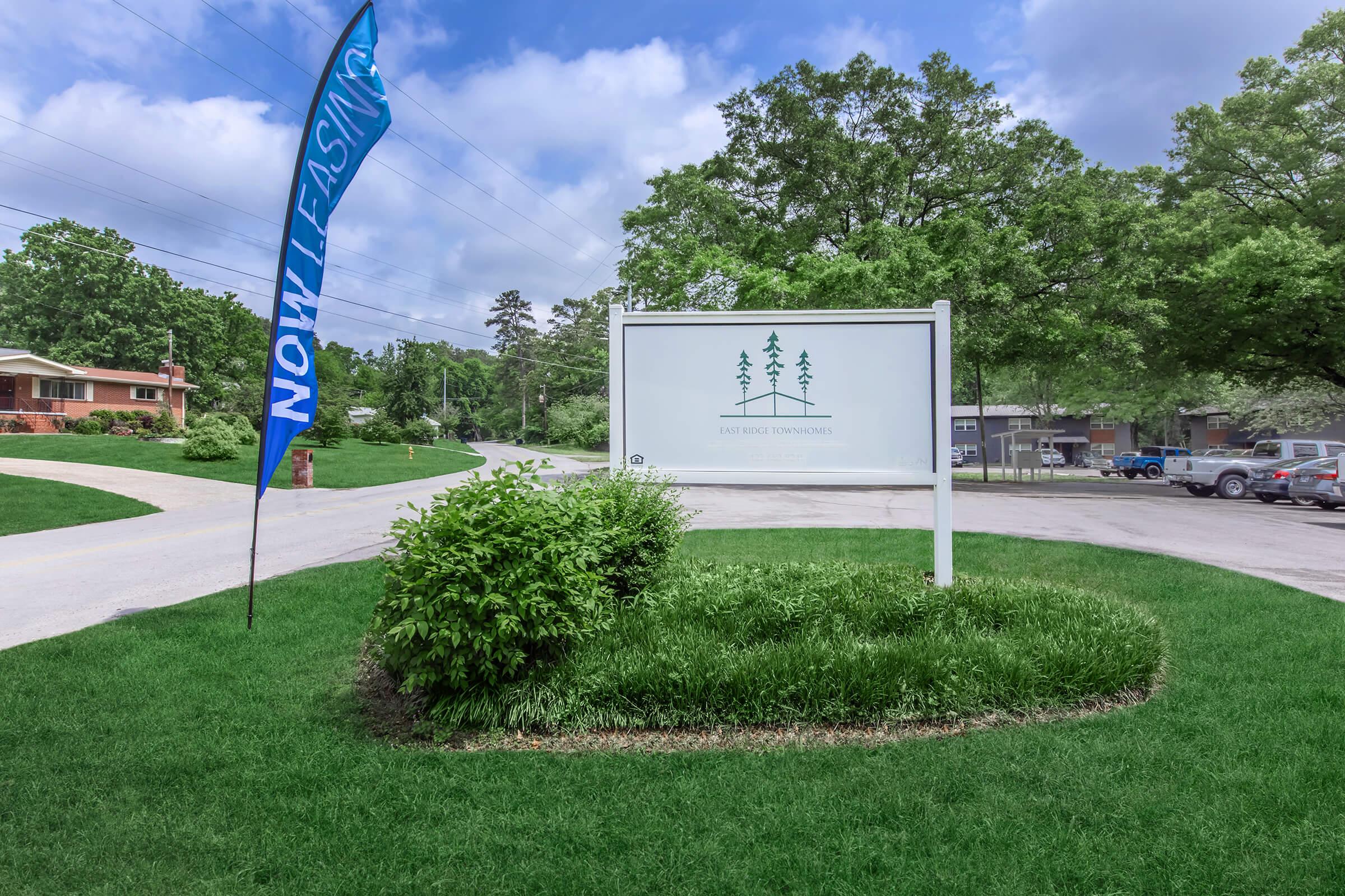Sign for "Riverbend Townhomes" surrounded by grassy landscaping, with a blue flag reading "NOW LEASING" nearby. In the background, residential buildings and trees are visible under a partly cloudy sky.