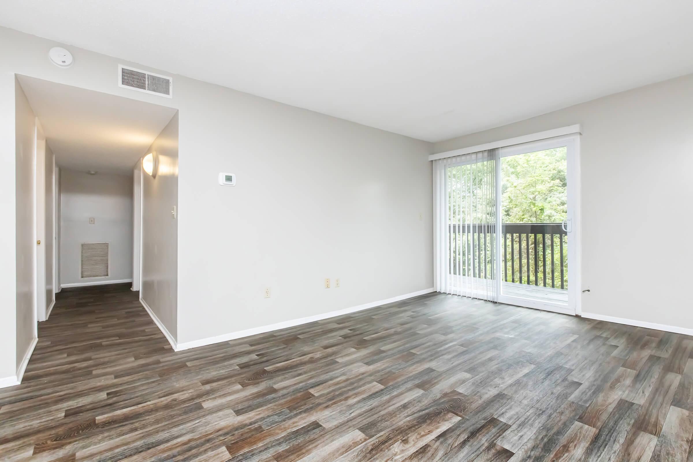Spacious and bright living area featuring wooden floors, neutral-colored walls, and a sliding glass door leading to a balcony with views of greenery. A hallway is visible to the left, enhancing the open and airy feel of the space. Natural light floods the room, creating a welcoming atmosphere.
