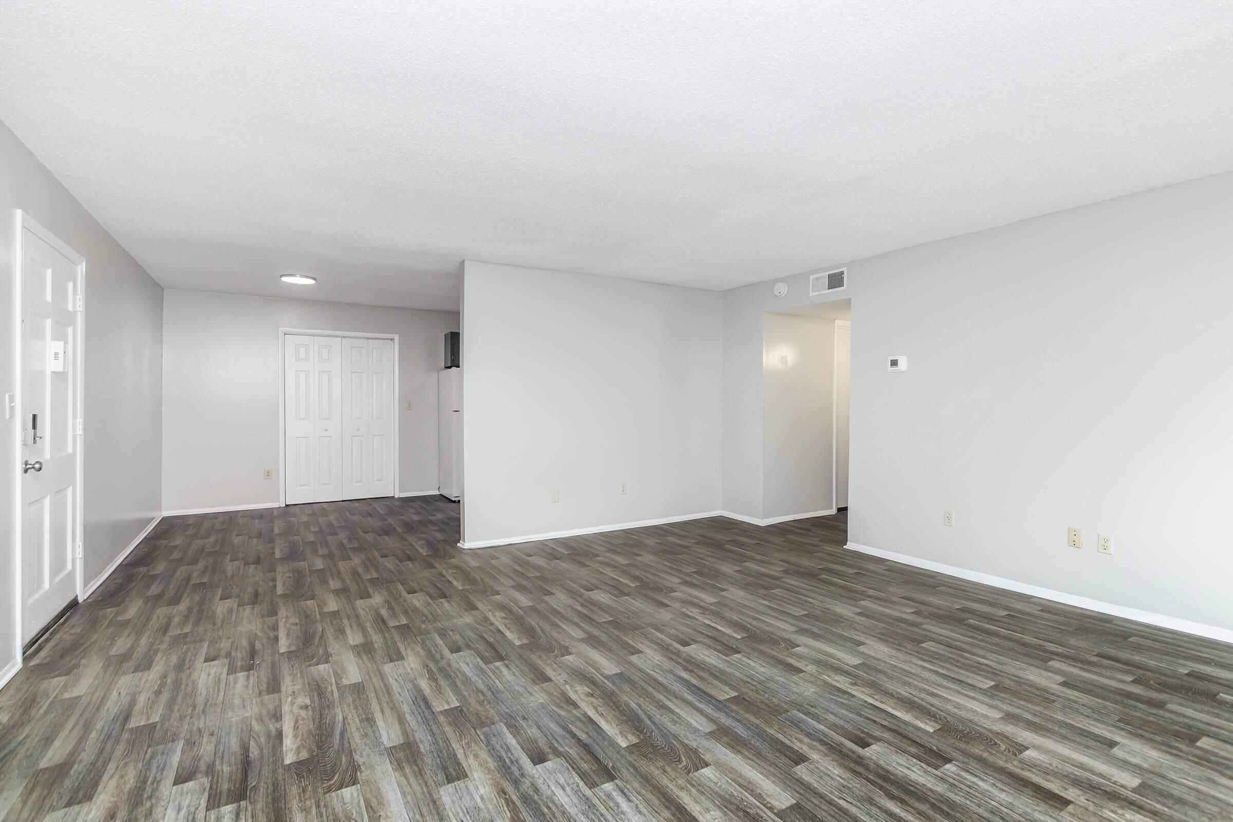 Spacious interior of an empty room featuring light-colored walls and modern laminate flooring. The room has a doorway on the left leading to an entrance, and a small hallway visible in the background. Natural light fills the space, creating an open and inviting atmosphere.