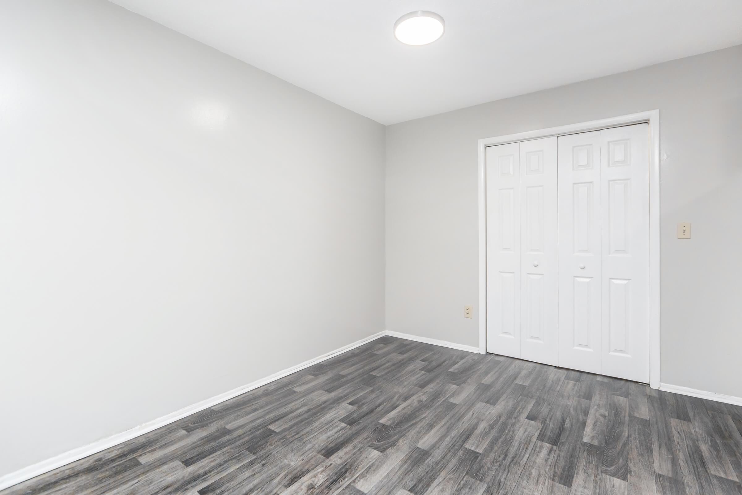 A spacious, empty room with light gray walls and a smooth wooden floor. There is a ceiling light fixture providing illumination, and a set of double doors leading to a closet or another area. The overall appearance is clean and minimalist, ideal for various purposes such as a bedroom or office.