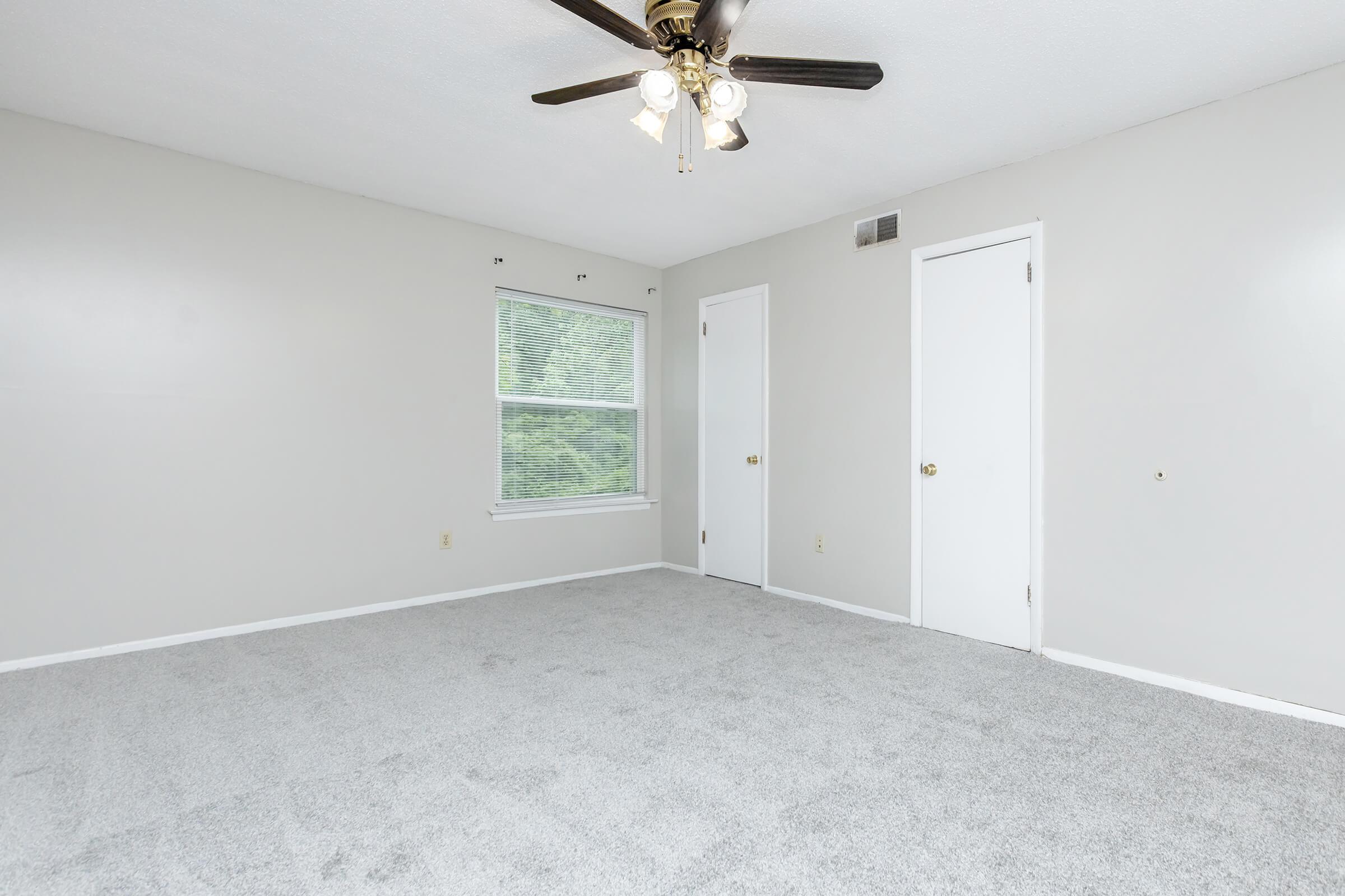 Empty room with light gray carpet, neutral gray walls, and a white ceiling fan with lights. A window allows natural light, and there are two closed white doors along the walls. The overall space appears bright and spacious, suitable for various uses.